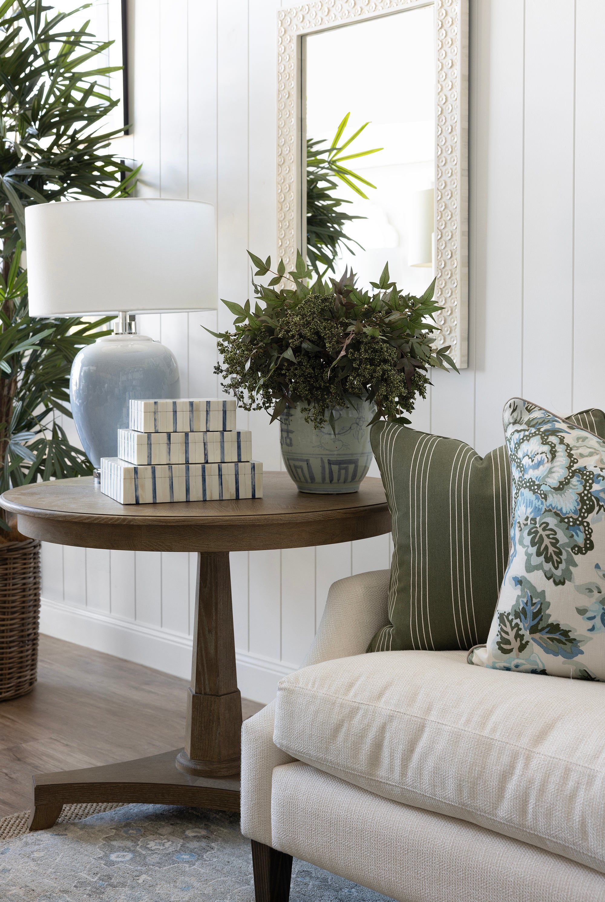 A styled interior corner featuring a round wooden side table with a white lamp, stacked decorative boxes, and a potted green plant, next to a cream sofa with green and floral throw pillows.