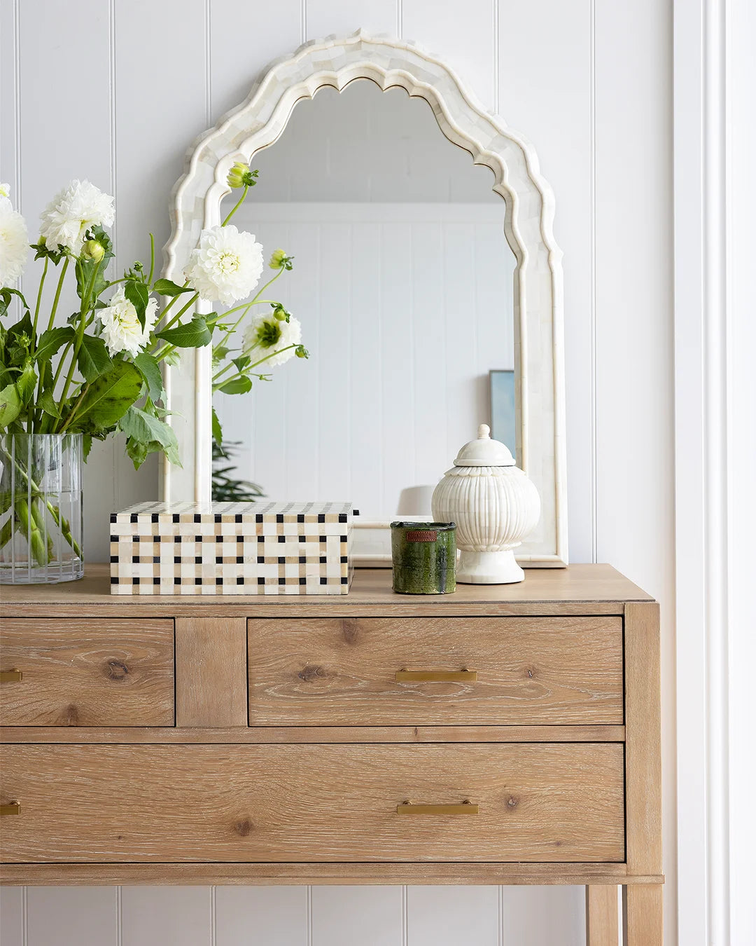 Bone scalloped arch mirror sitting on oak console, leaning against a white vj wall. Vase of white flowers, checkered box, a green candle and decorative bone jar sit on top of the console.
