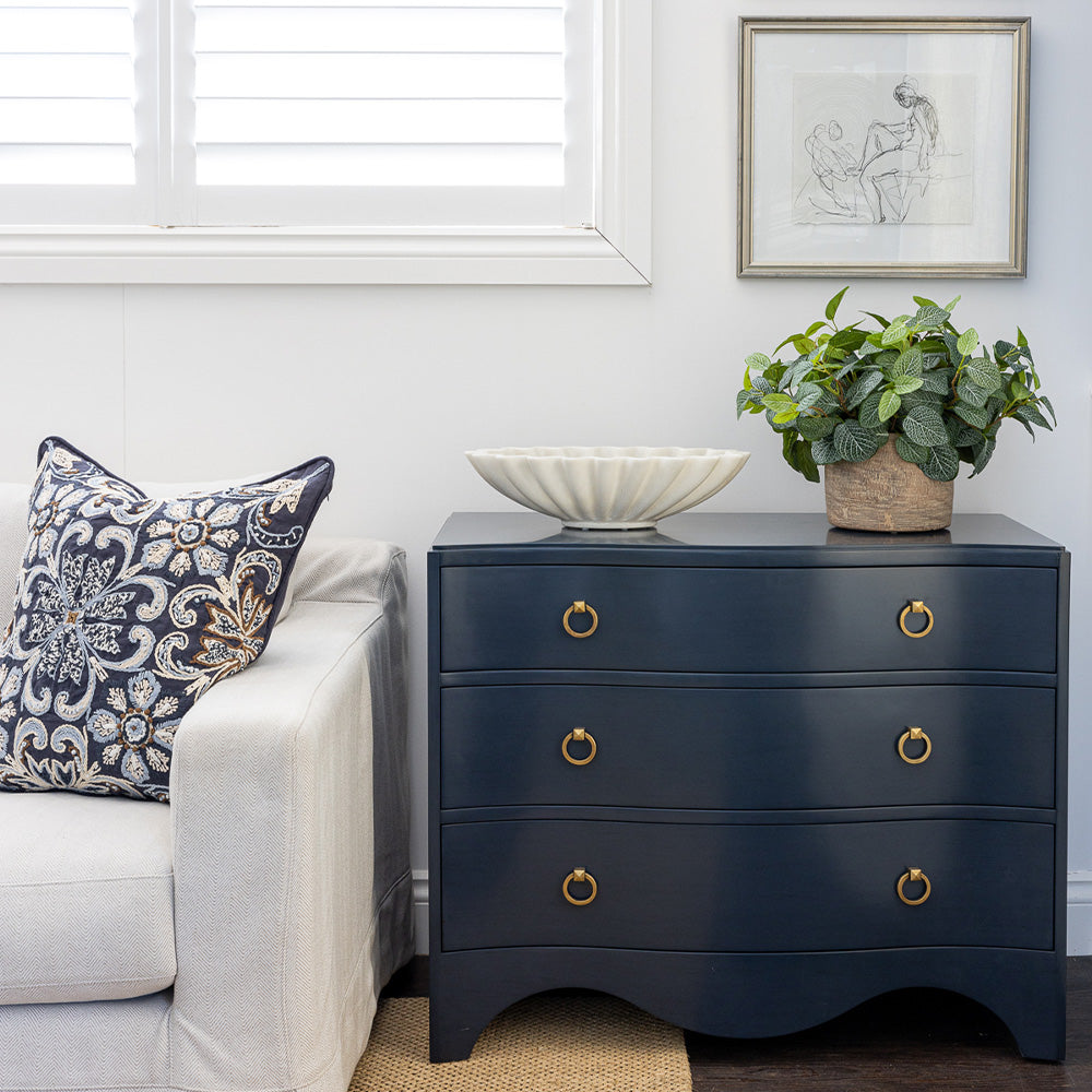 Image of Navy Claire Chest styled next to a neutral sofa with a navy embroidered cushion. The chest is styled with an oval ruffled marble bowl and faux fittonia greenery on top, figure study artwork hanging on wall above.