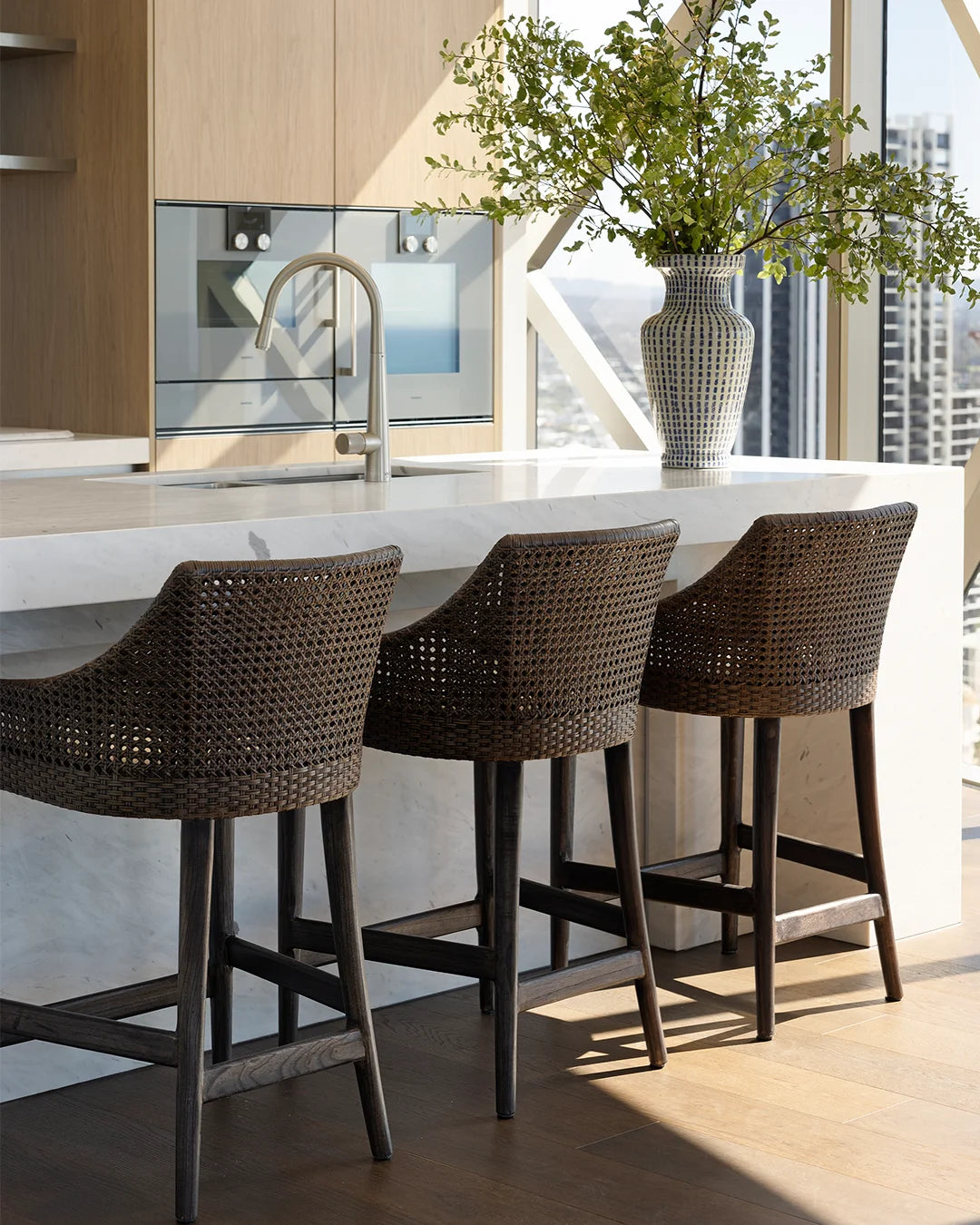 Three dark brown woven wicker bar stools with dark wooden legs are arranged along a stone kitchen island in a modern kitchen setting with a vase of greenery nearby.