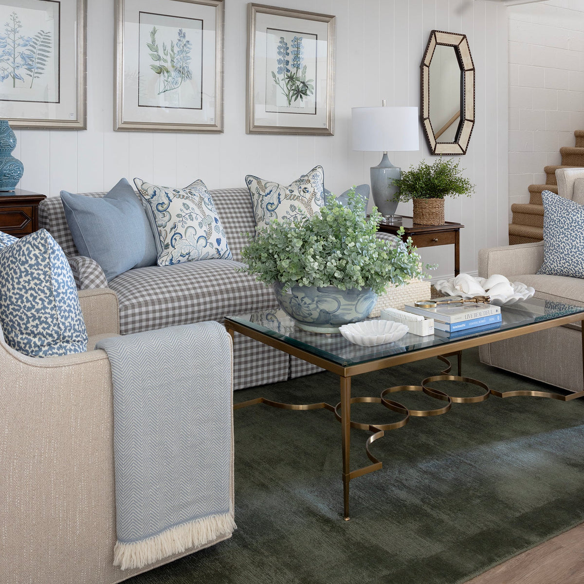 A styled living room interior featuring a beige armchair, a blue-and-white checkered sofa with blue accent pillows, a glass-top coffee table with gold legs, framed botanical prints on the wall, and a white ceiling with exposed beams.