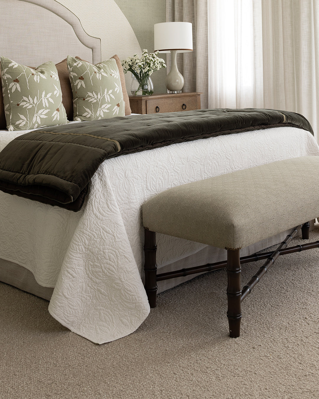faux bamboo bench at end of bed, Bed with white bedspread, green comforter, green embroidered cushions with caramel cushions behind, custom neutral upholstered bedhead, oak bedside table with an almond coloured lamp and vase of small white flowers.