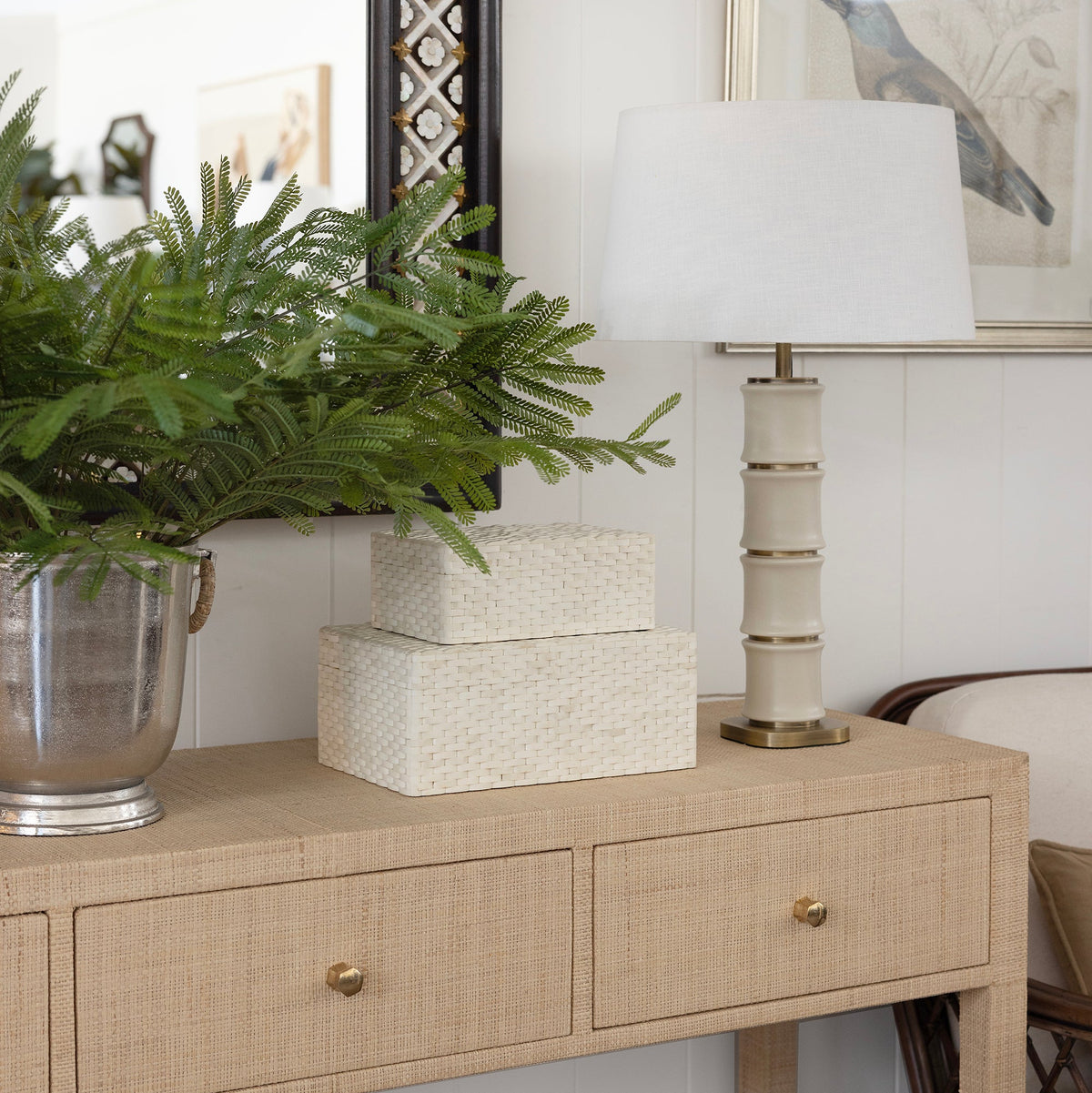 A raffia woven console table in a styled room setting, decorated with a green fern in a silver pot, two stacked white textured boxes, and a cream-colored table lamp with a white shade.