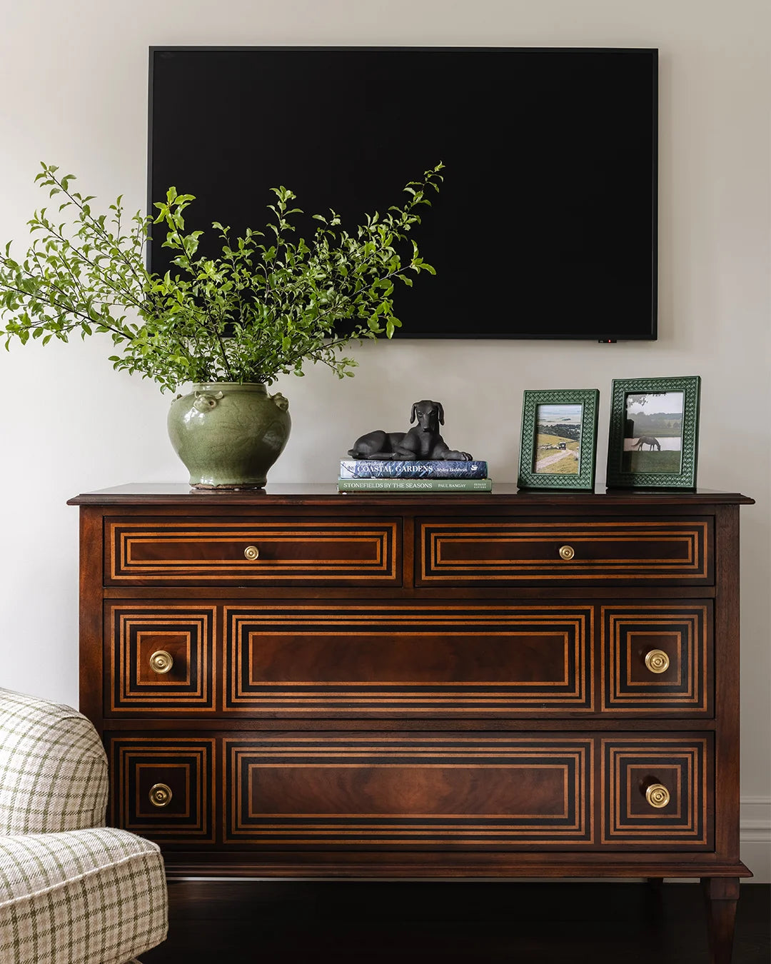 Chest styled with greenery, books with dog sculpture and green leather frames. TV mounted above chest.