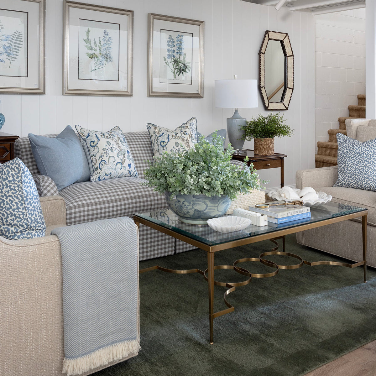 A styled living room interior featuring a beige armchair, a blue-and-white checkered sofa with blue accent pillows, a glass-top coffee table with gold legs, framed botanical prints on the wall, and a white ceiling with exposed beams.