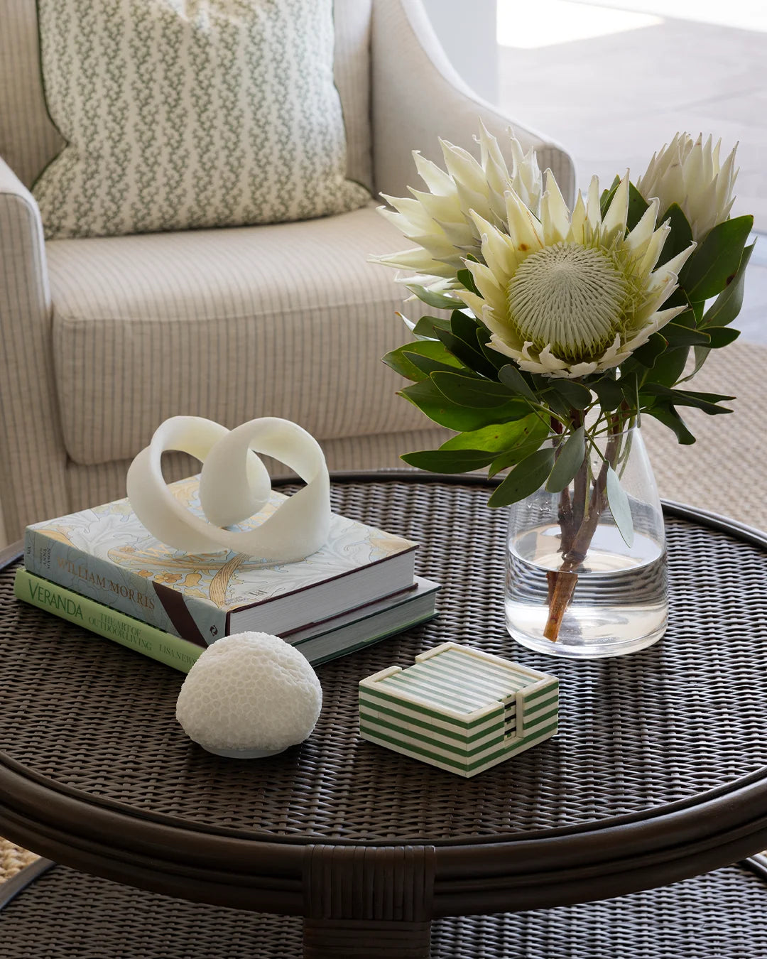 Rattan round coffee table styled with green bone striped coasters, books and object and a vase of cream proteas. Upholstered neutral stripe chair with green patterned cushion in background.