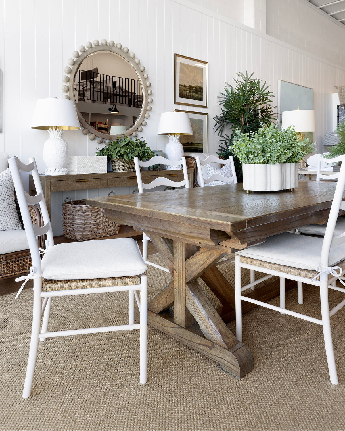A rustic wooden dining table with a trestle base, surrounded by white chairs with white seat cushions and woven backs, set in a bright dining room with a console table, lamps, and plants.