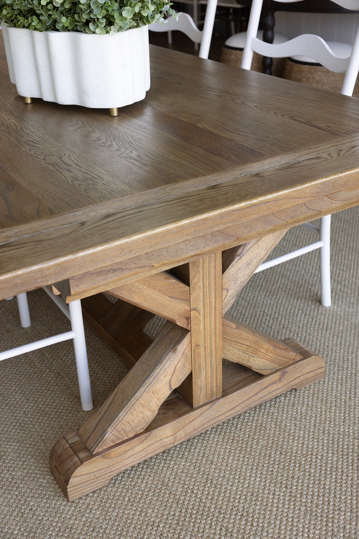 A rustic wooden dining table with a thick rectangular top and a trestle-style base, shown in a real interior setting with white chairs and a white planter visible around it.