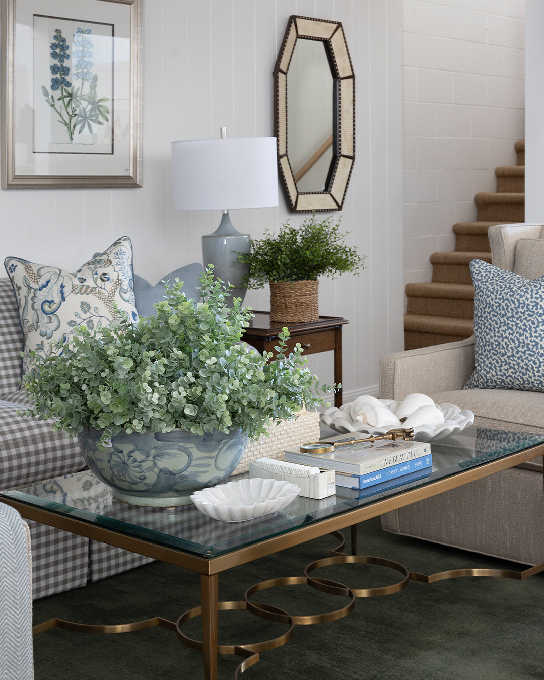 A styled living room interior featuring a glass-top coffee table with a gold metal frame, a blue ceramic planter with green foliage, a gray and white checkered sofa with blue patterned pillows, and a beige armchair in a bright, modern room with white ceiling beams and a staircase.