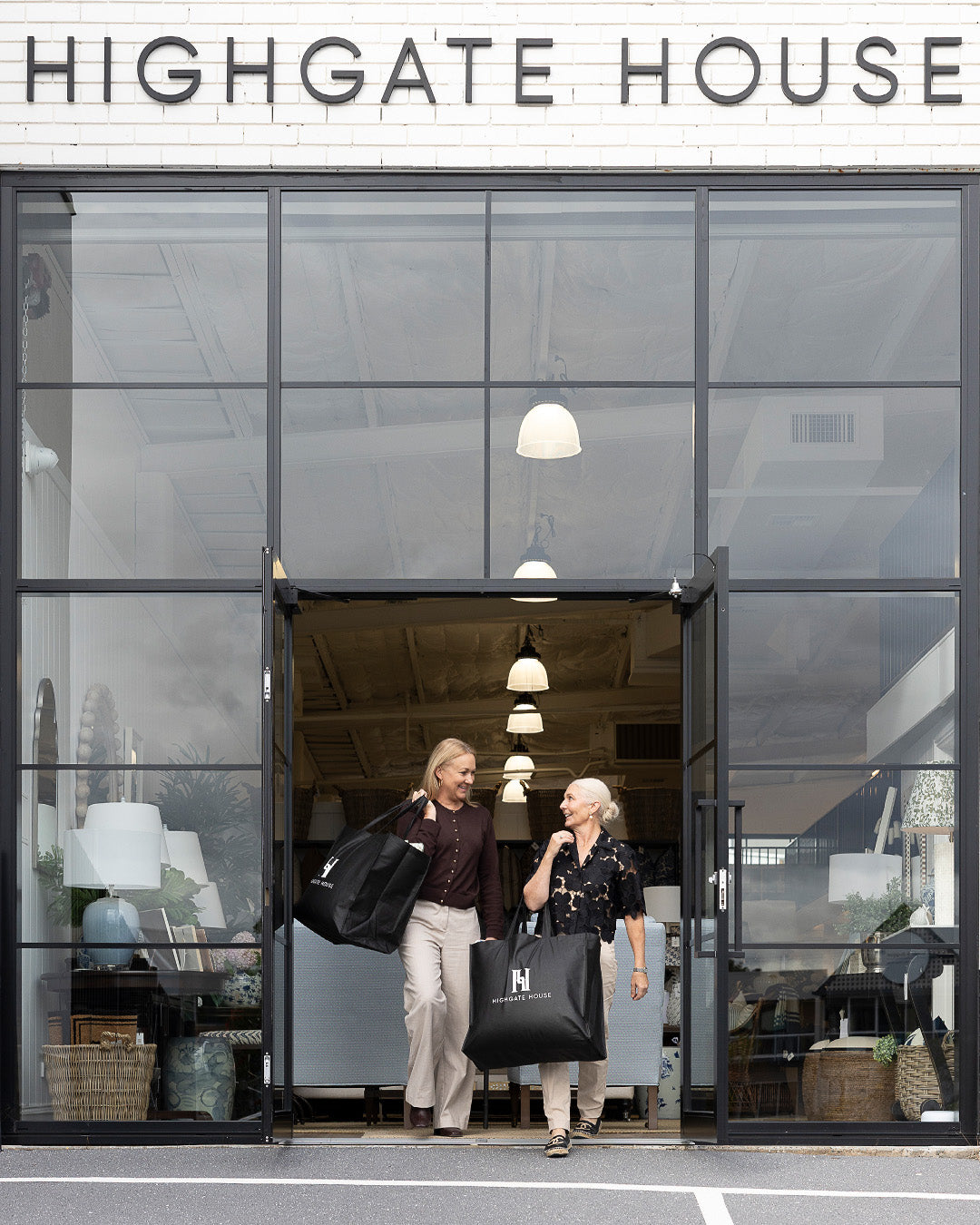 Two women walk out of a modern storefront with large glass windows and black frames. The sign above reads HIGHGATE HOUSE.