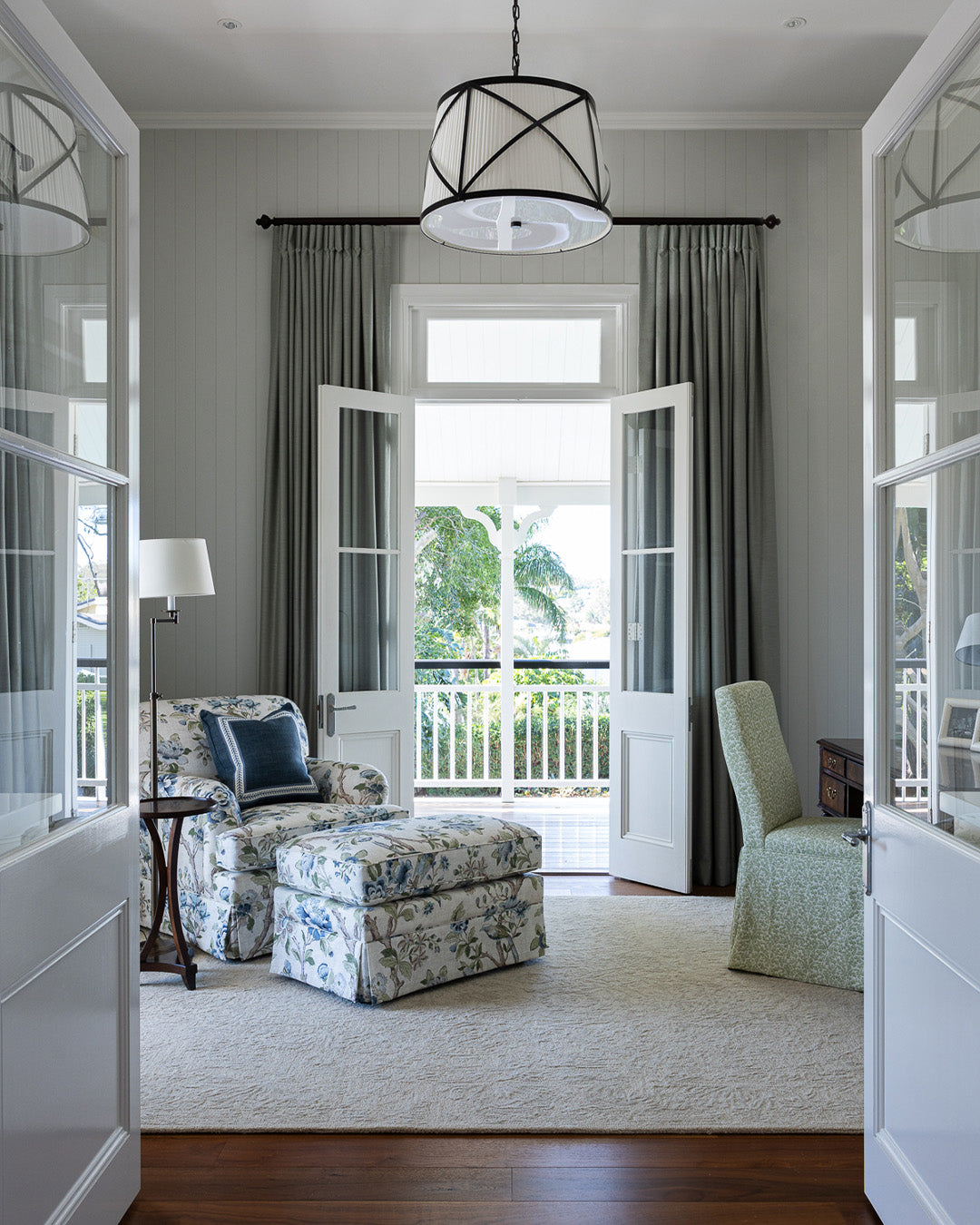 A bright, elegant room viewed through an open doorway, featuring gray curtains, a black metal pendant light, a floral armchair with matching ottoman, a light green upholstered chair, and a large cream rug.