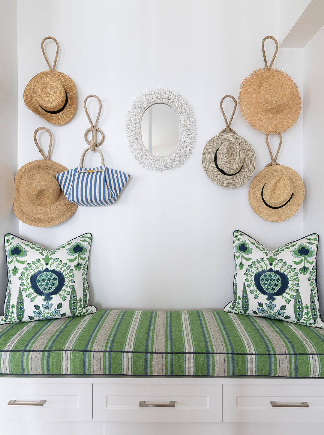 A styled entryway bench with a green and white striped cushion, two matching patterned throw pillows, and a white textured oval mirror on a white wall above. Several straw hats and a blue-and-white striped handbag hang from rope loops on the wall.