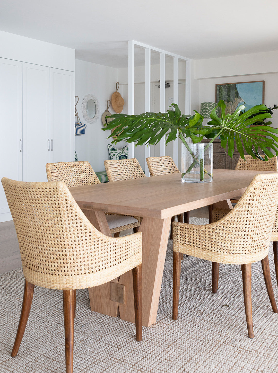 A mid-century modern dining table with a light oak wood top and thick wooden legs, surrounded by six woven cane chairs, set on a textured rug in a bright, modern dining room.
