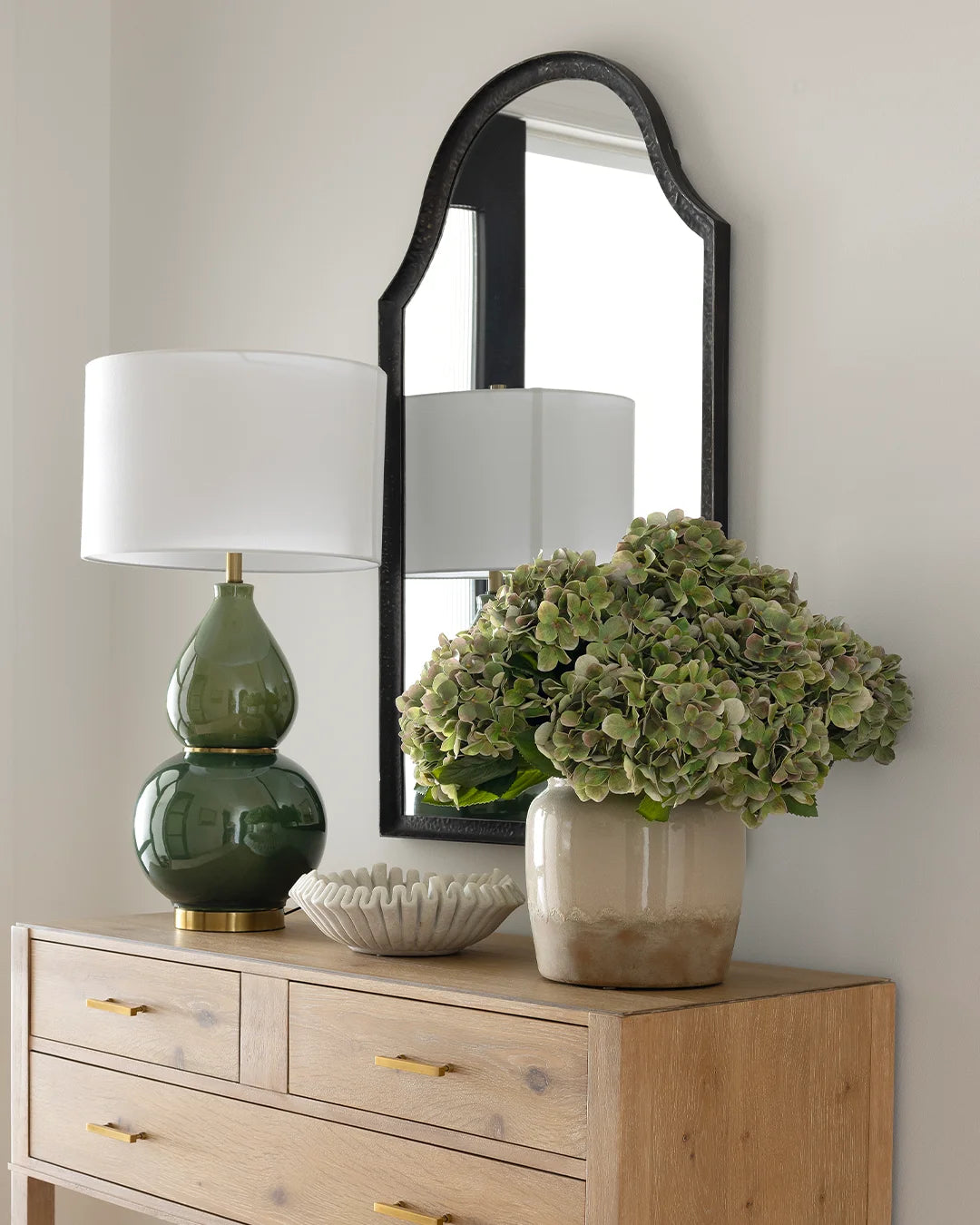 Polly chest with green lamp, marble bowl and vase of faux green hydrangeas, styled against white wall with black frame mirror.