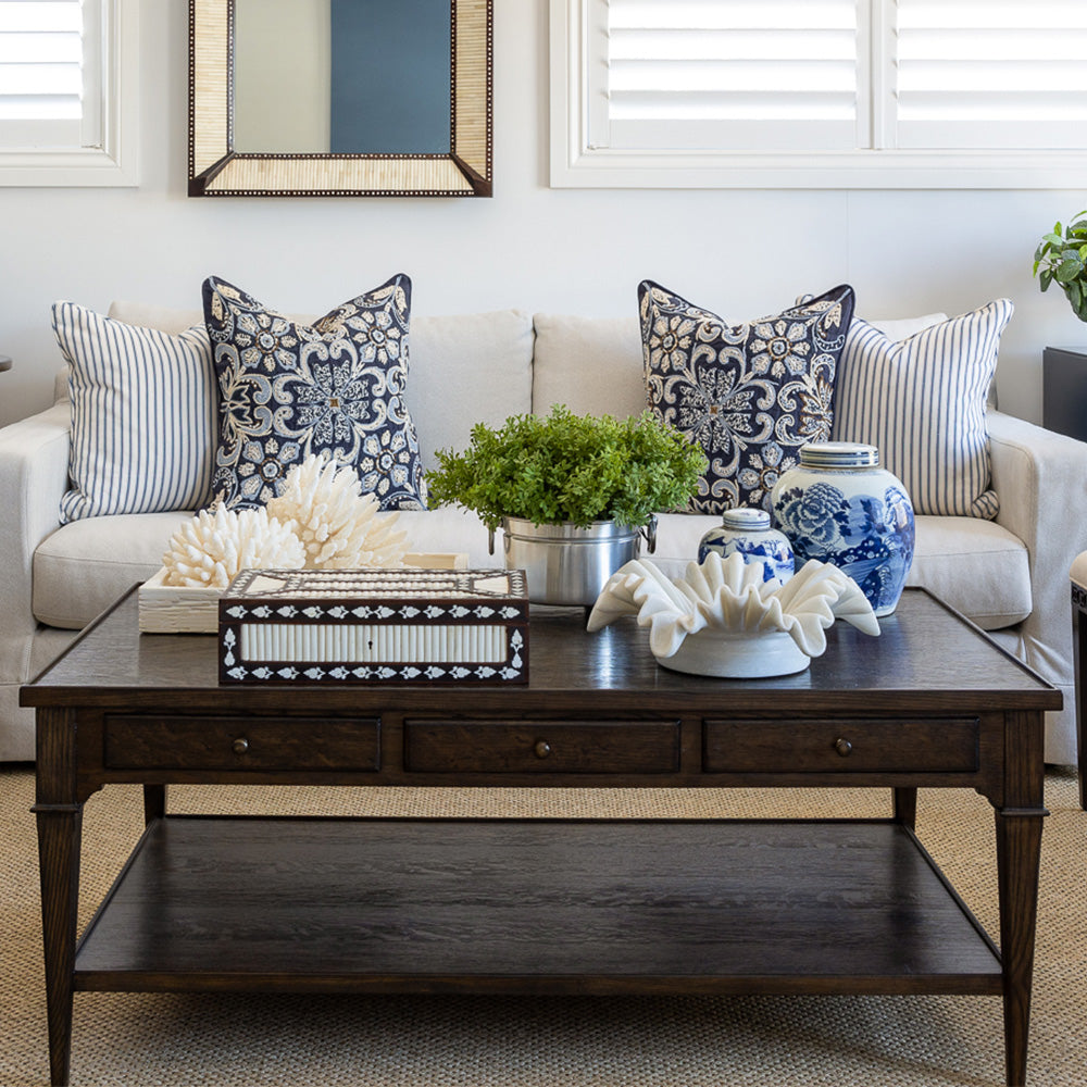 Huxley coffee table styled in front of a neutral sofa with navy embroidered and navy and white striped cushions. The coffee table is styled with a wooden bone inlay box, a ruffled marble bowl, two small blue and white ginger jars, a faux greenery arrangement and a carved bone inlay tray with decorative coral.