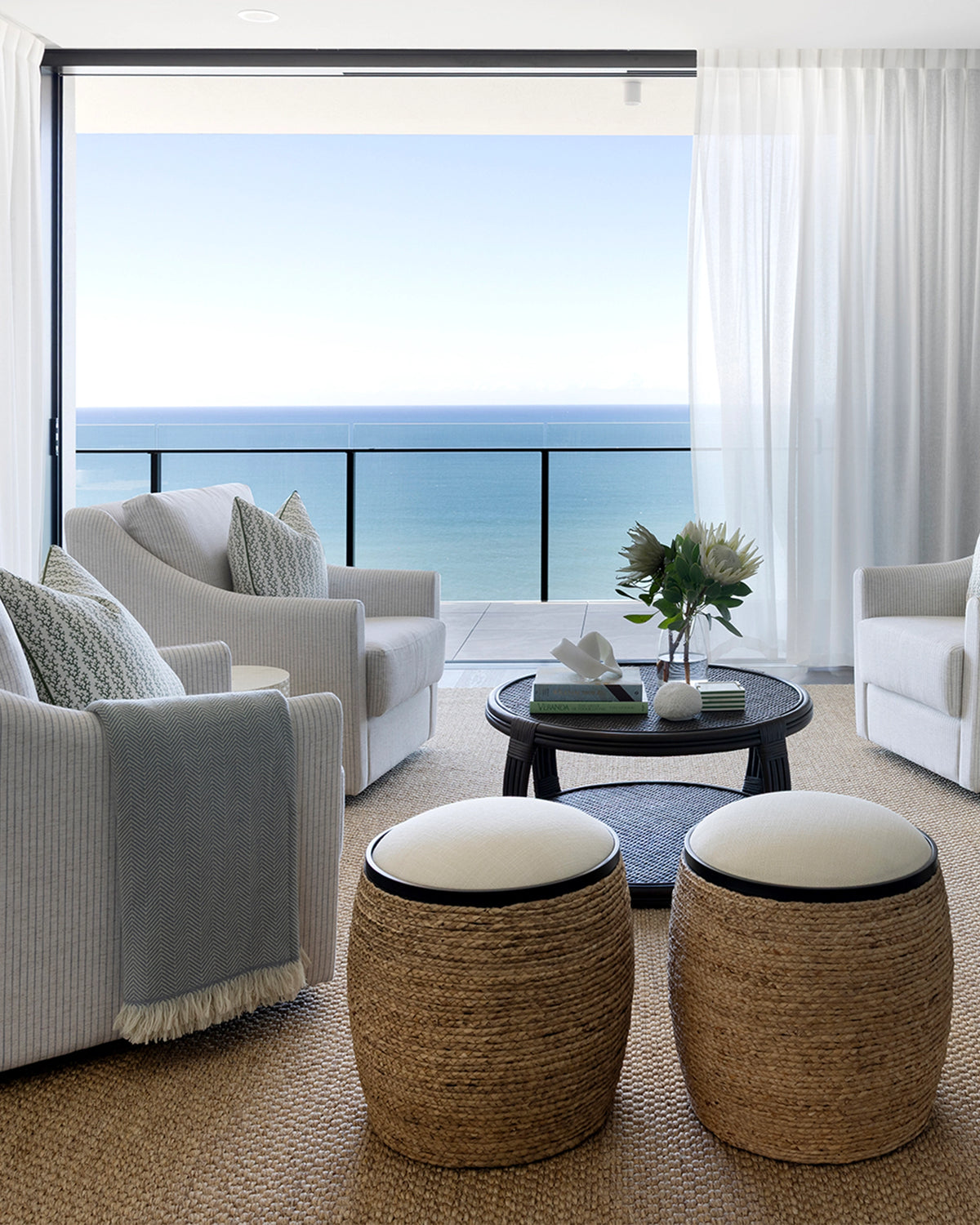 A bright, modern living room with a large ocean view from floor-to-ceiling windows. The room features light gray and white upholstered armchairs, two round woven ottomans with cream cushions, a dark round coffee table with flowers, and a textured beige rug.