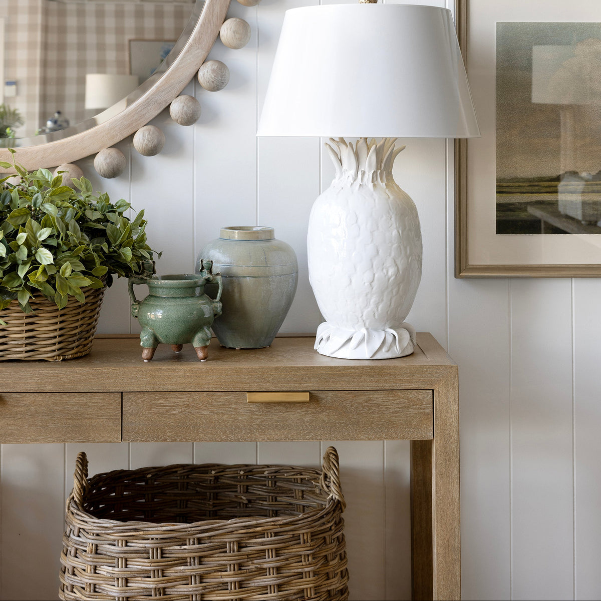 A white textured ceramic table lamp with a tapered shade sits on a light wood console table against a white shiplap wall, styled with a green plant, ceramic vases, a round beaded mirror, and framed landscape prints.