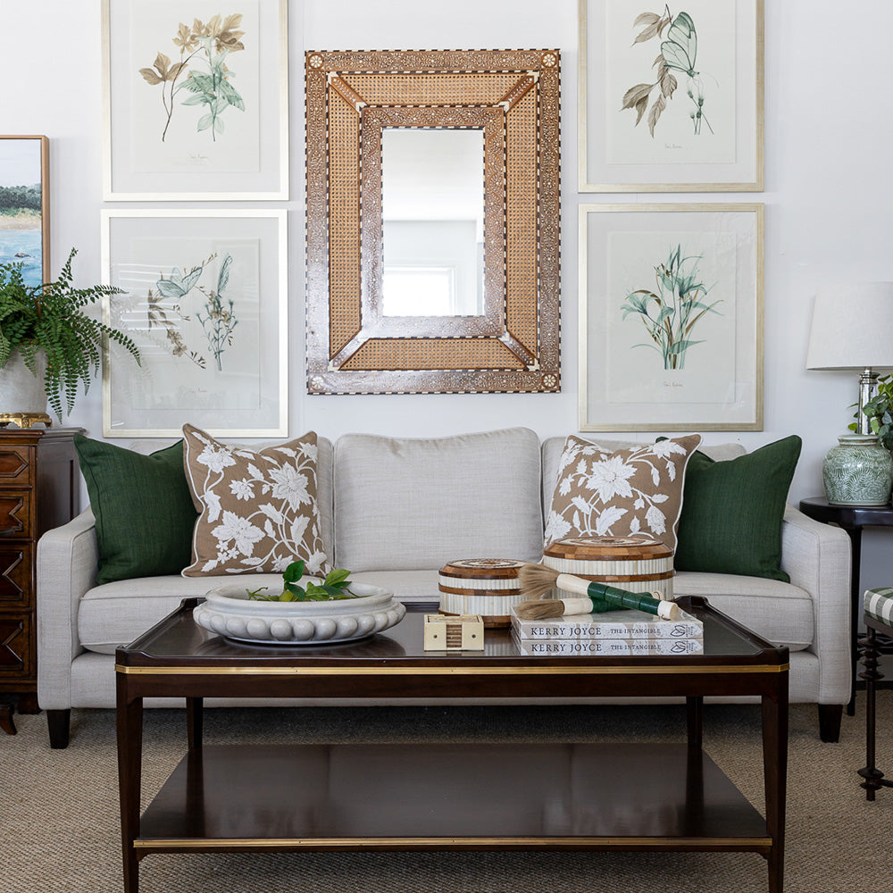 Image of maddox coffee table styled in front of a neutral sofa with green and caramel cushions. The coffee table is styled on top with a large marble bowl filled with limes, a set of cream coasters, teak and bone inlay round boxes and a stack of books topped with ornamental paint brushes. Behind the coffee table is a large rattan and bone inlay decorative mirror on the wall.