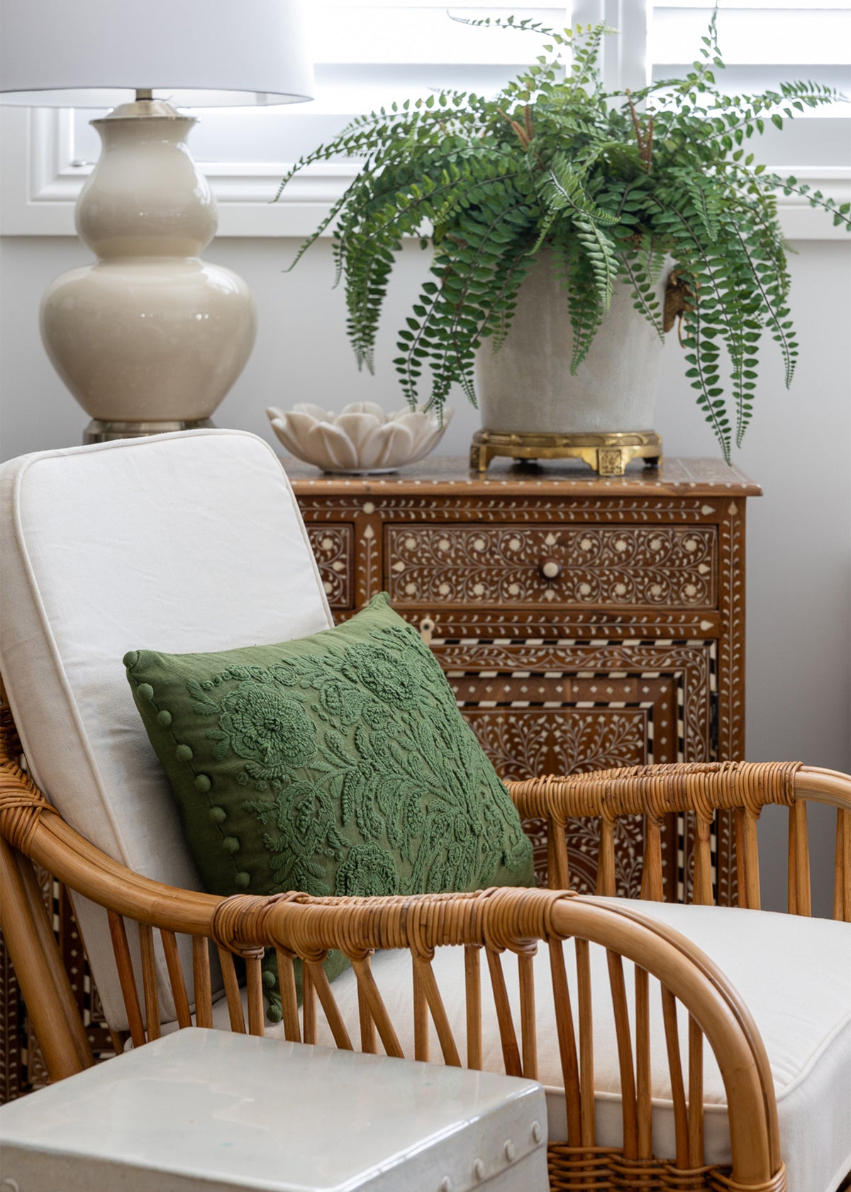 Mara cabinet styled in a living space. With a Rattan chair in front. The cabinet is styled with a large faux fern arrangement a small marble lotus bowl and a cream lamp.