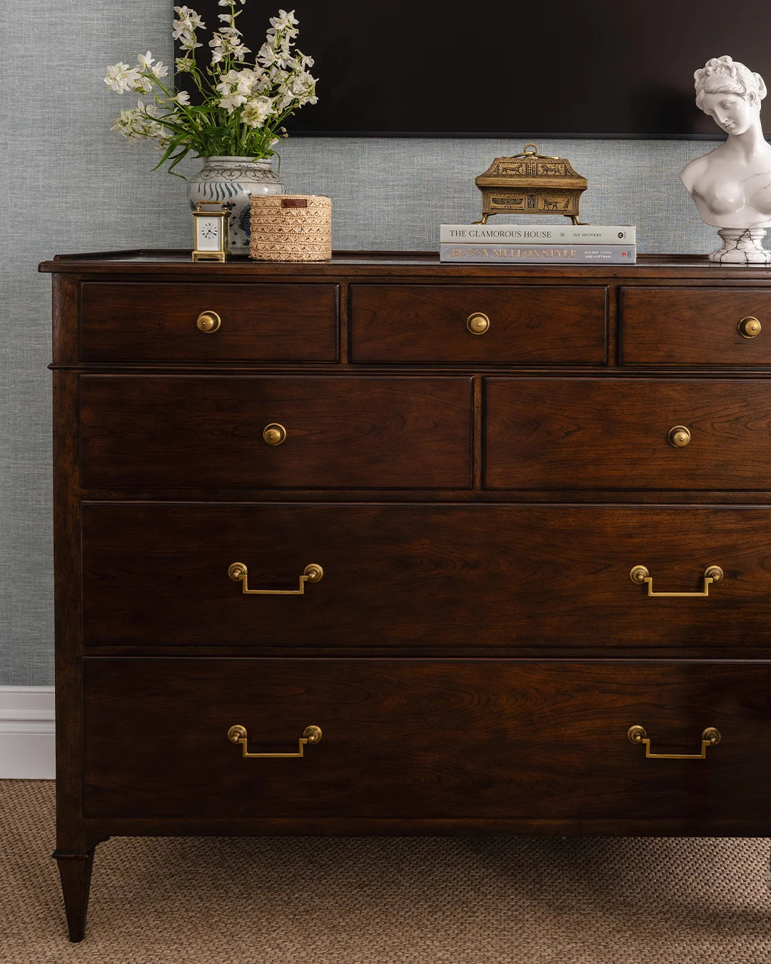 Chest styled with flowers, candle, and clock, a stack of books with brass box and a sculpture. Blue textured wallpaper behind with mounted TV above chest.