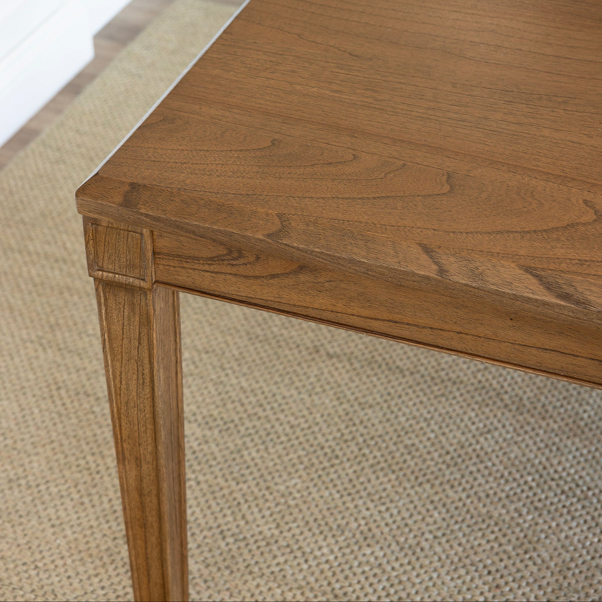 Close-up of a wooden dining table with a warm medium-brown finish and visible wood grain, shown in a home interior setting with a textured rug and white cabinetry in the background.