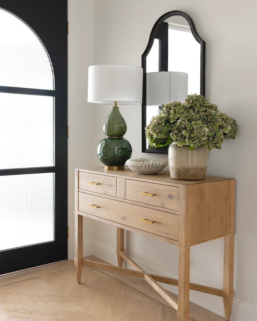 Oak console table in entry, styled against white wall, with a green lamp, marble bowl and vase of faux green hydrangeas. Black scalloped mirror on the back wall.