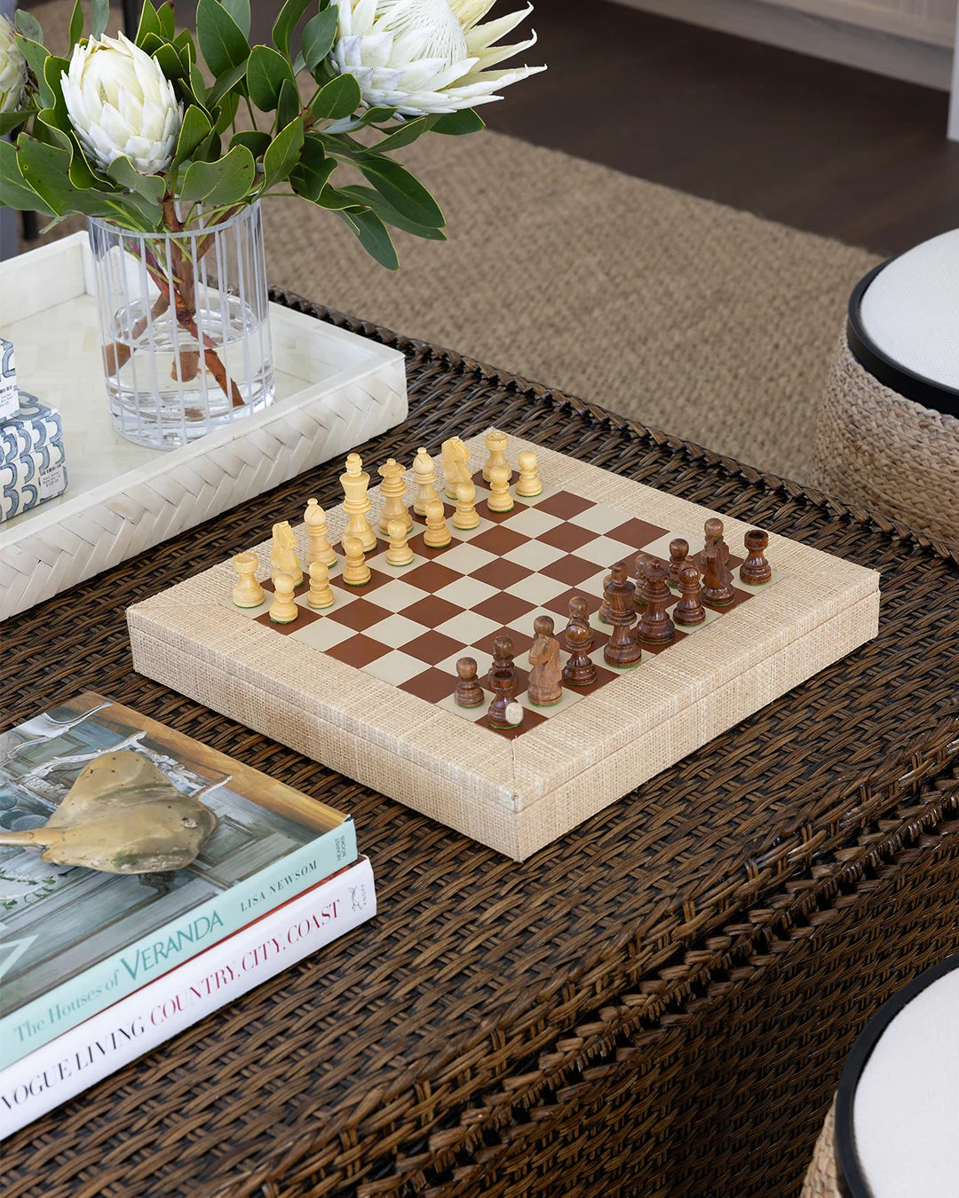 A wooden chess set with cream and dark brown pieces arranged on a checkered board, placed on a woven brown table. A glass vase with white flowers and a book are nearby in a styled indoor setting.