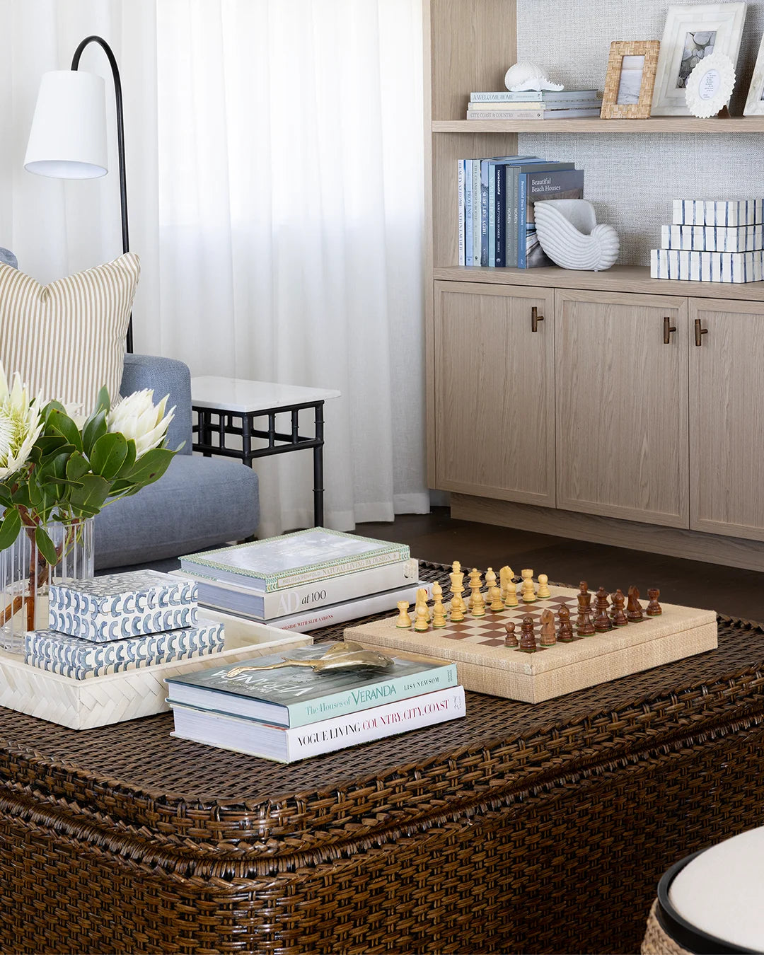 A dark brown woven rattan ottoman coffee table styled in a modern living room setting, holding a chess set, books, and decorative pillows.