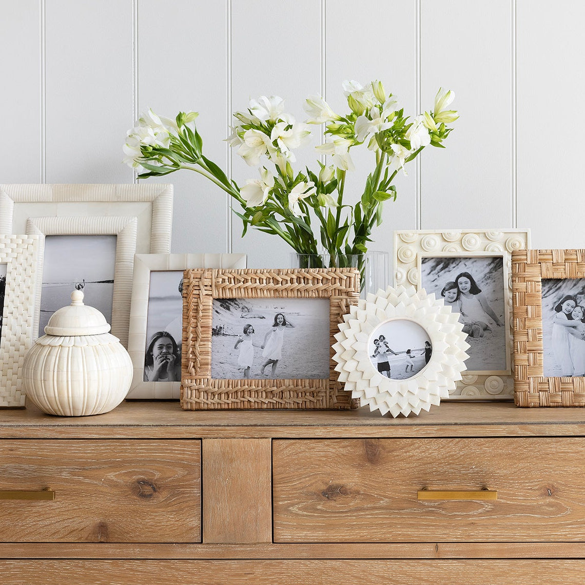 Wooden dresser with decorative items including framed photos and a vase of flowers against a white wall.