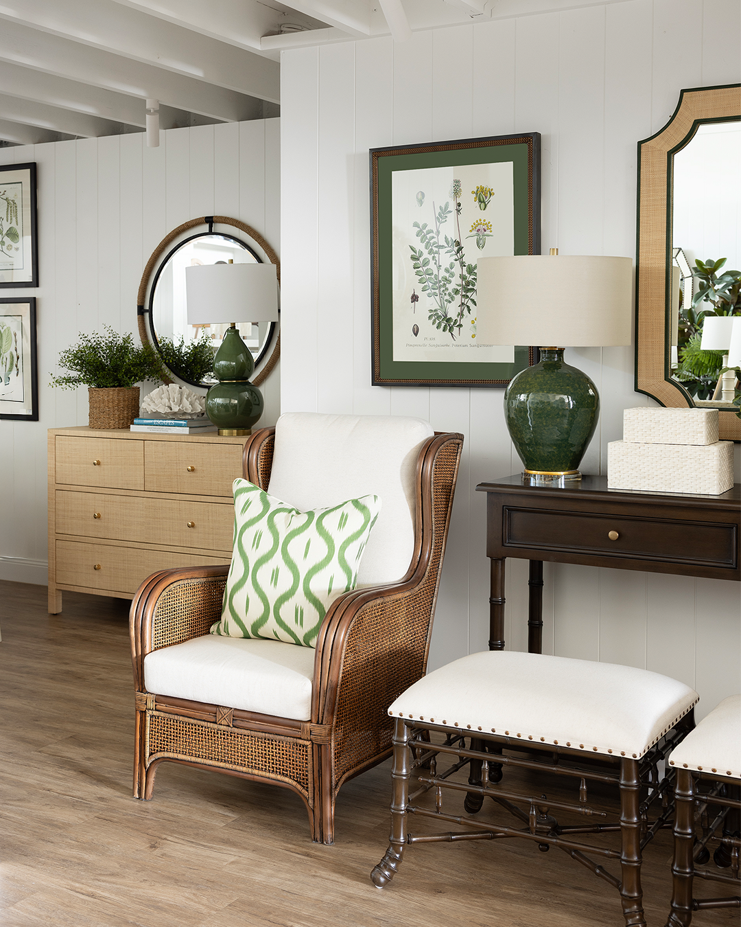 Rattan armchair with abstract green cushion sitting next to a console styled with an olive green lamp, bone boxes, and two ottomans in front.