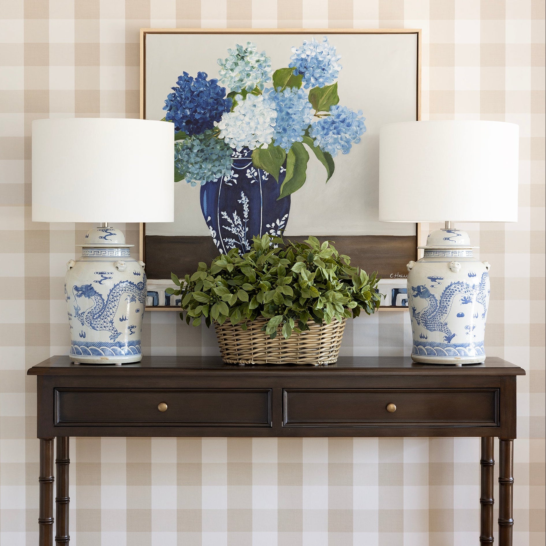 A dark brown wooden console table styled in a home entryway, flanked by two white table lamps with blue dragon-painted ceramic bases. A wicker basket filled with green foliage sits in the center, and a framed floral print hangs on the beige-and-white checkered wallpapered wall behind it.