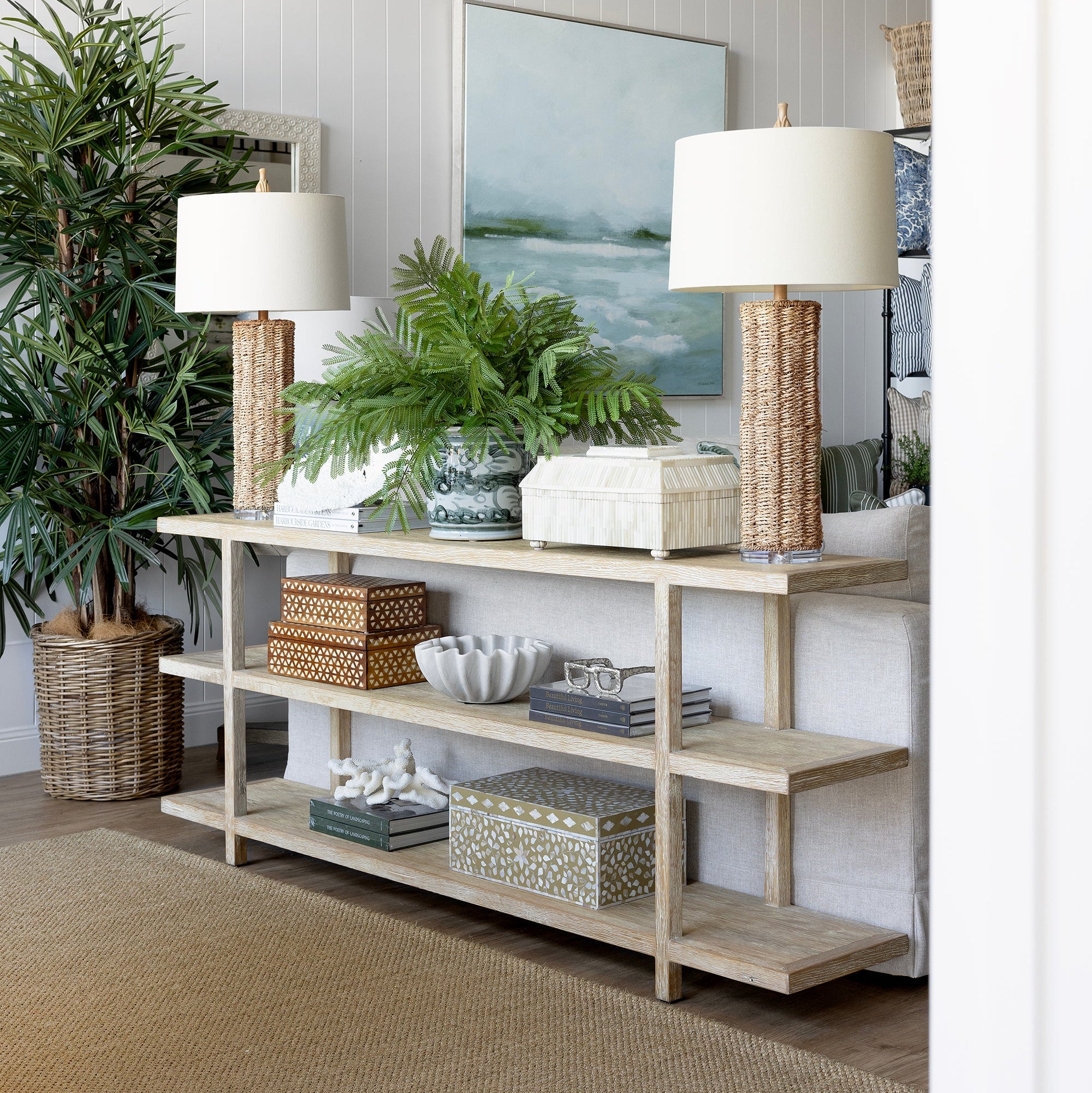 A light natural wood console table styled in a bright, modern living room with white paneled walls, two table lamps, potted green plants, and decorative baskets and boxes.