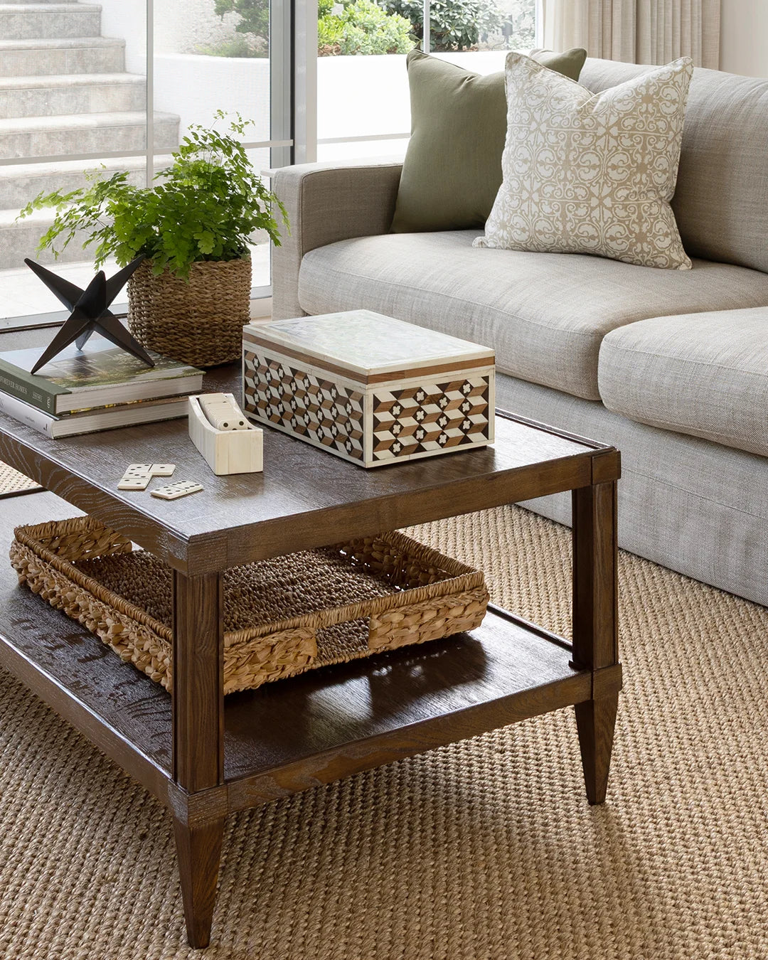 A dark brown wooden coffee table with a lower shelf, styled in a bright living room with a woven basket, books, and a potted plant.