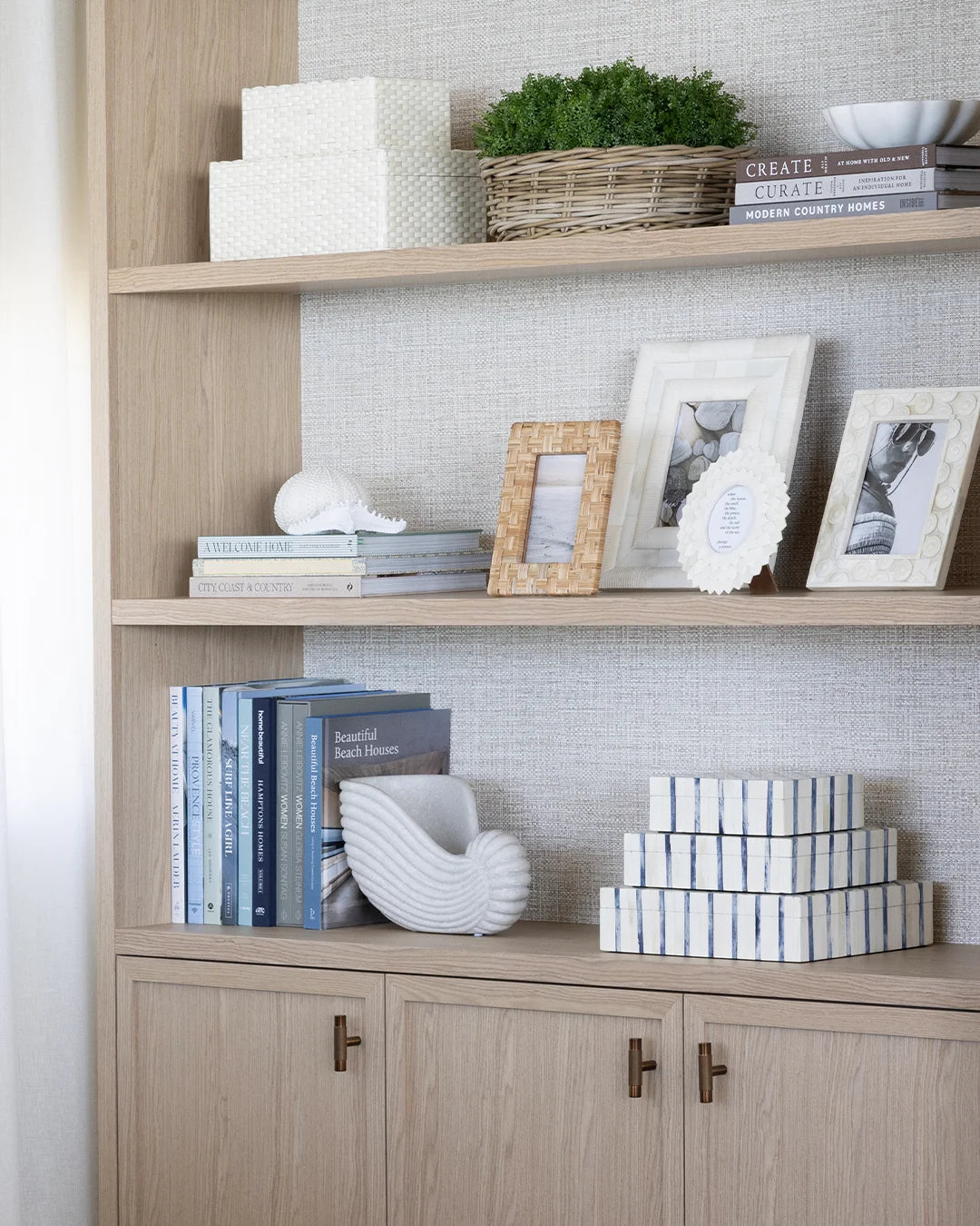 A light wood built-in shelving unit with a textured light gray wall behind it, styled with books, framed photos, a basket planter, and stacked white boxes.