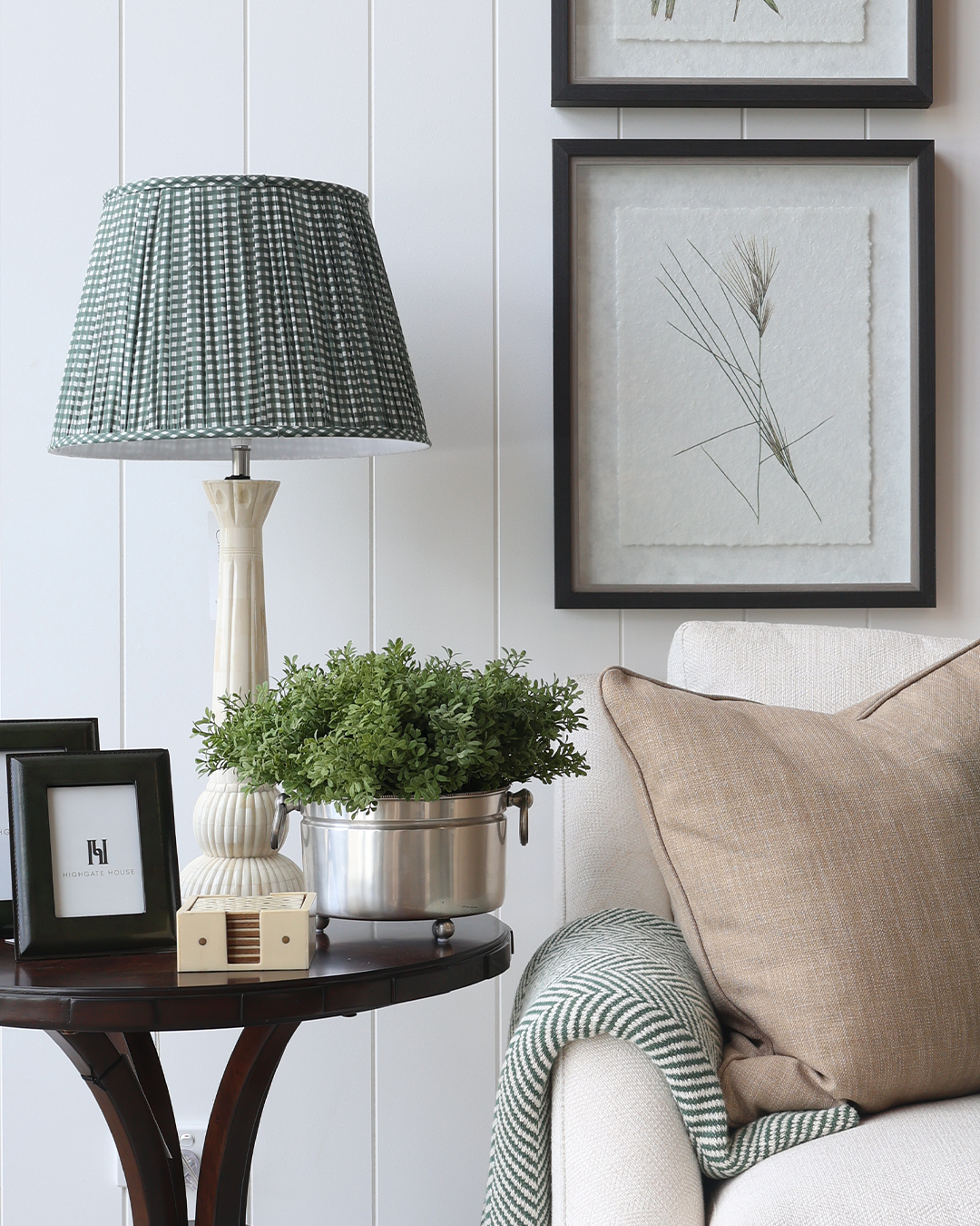 A styled interior scene showing a table lamp with a textured green and white patterned shade and a cream base on a dark wood side table. A potted green plant, framed botanical wall art, and a beige sofa cushion are visible in a real room setting.