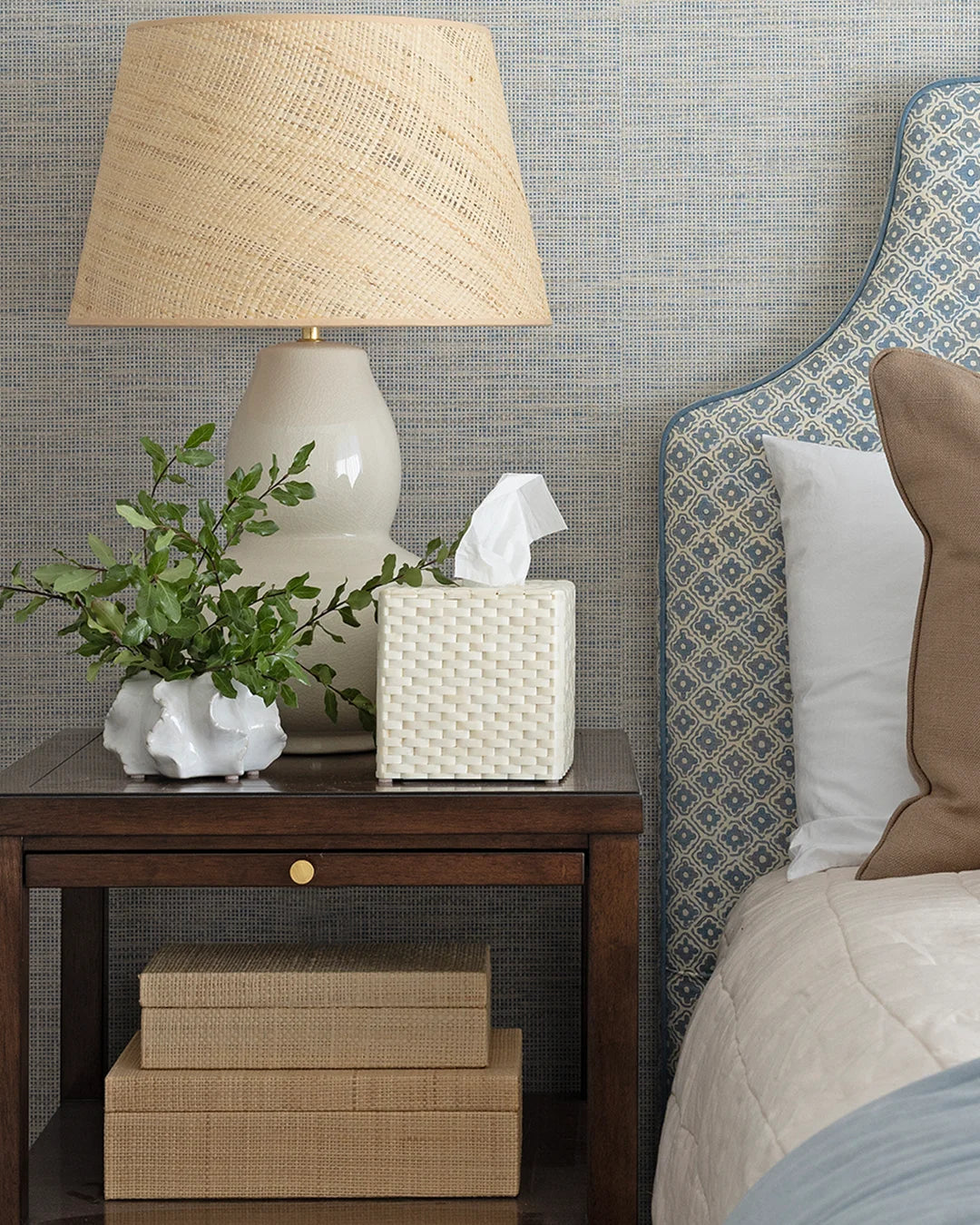 A styled bedroom corner featuring a dark wood nightstand with a white ceramic table lamp, a white tissue box cover, a small white planter with greenery, and stacked woven boxes, next to a bed with a blue patterned headboard.