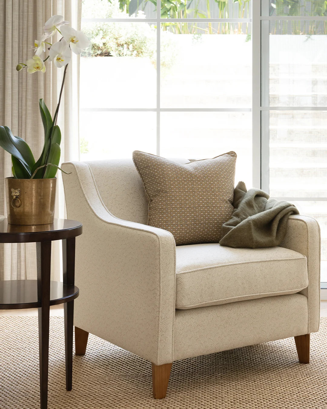 Olive throw on upholstered chair with beige patterned cushion, dark timber side table and orchid in pot.