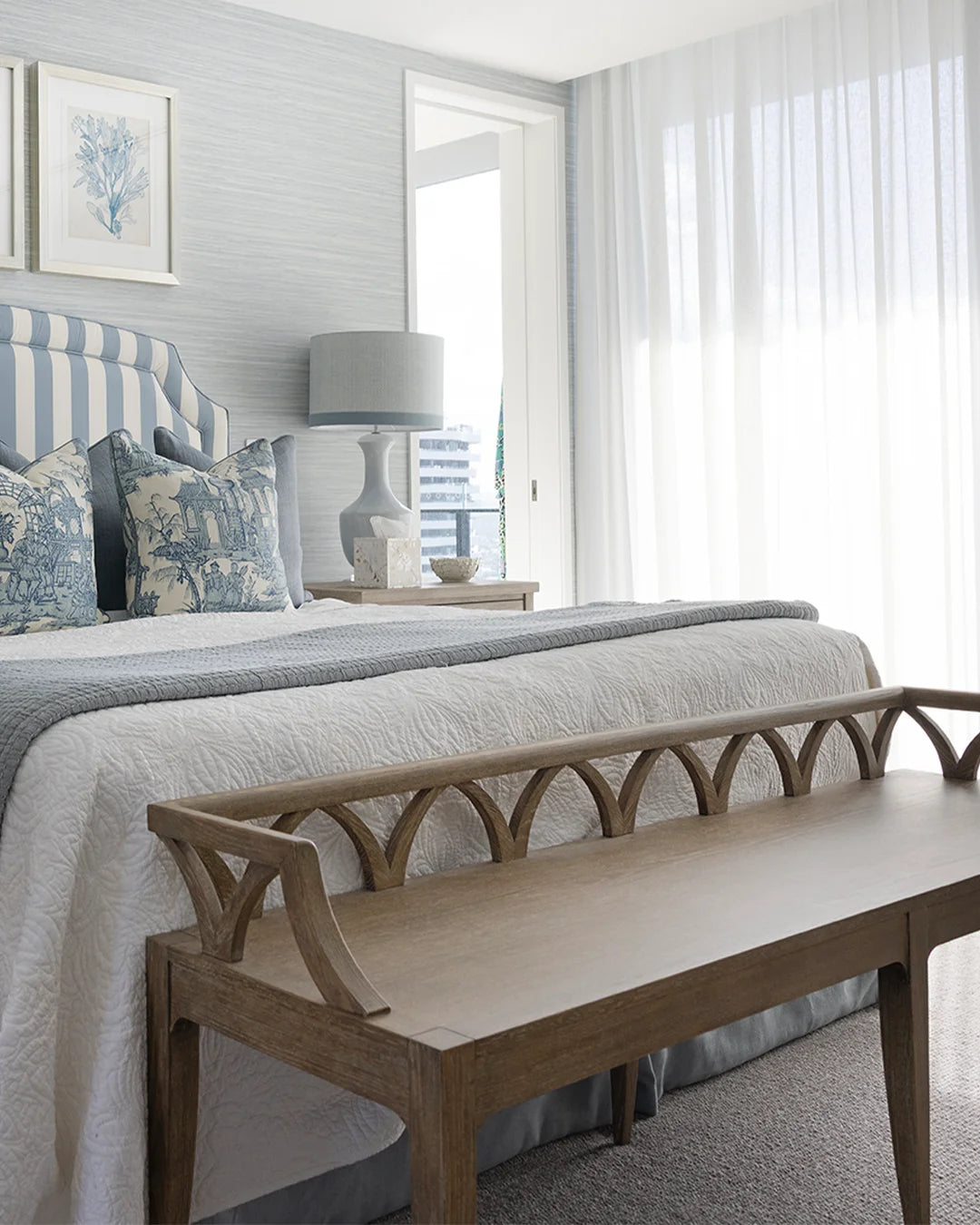 A styled bedroom interior featuring a wooden bench at the foot of a bed with white and blue bedding, striped pillows, a striped headboard, a bedside table with a lamp, and sheer white curtains by a bright window.