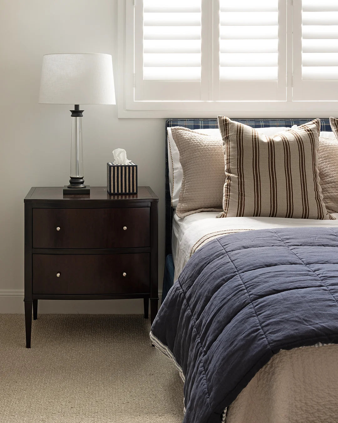 Dark timber bedside styled with lamp and striped navy tissue box sitting beside bed with chocolate and navy bedding.