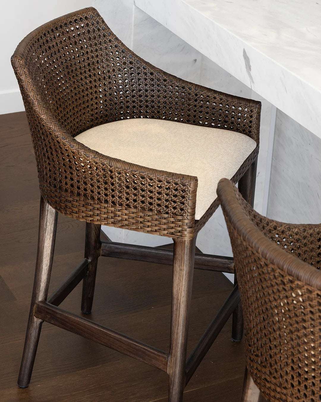 A brown woven rattan barstool with a dark wood frame and a light beige seat cushion, shown in a dining or bar setting next to a white marble counter.