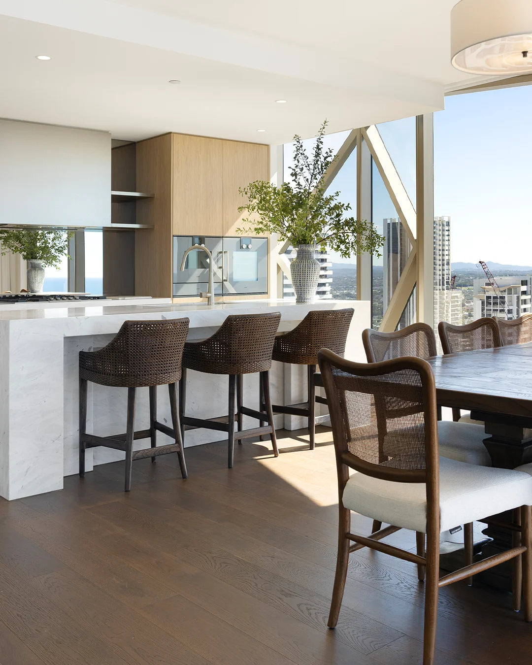 A modern dining and kitchen interior with a white marble island, four dark woven bar stools, a wooden dining table, and large windows overlooking a city skyline.