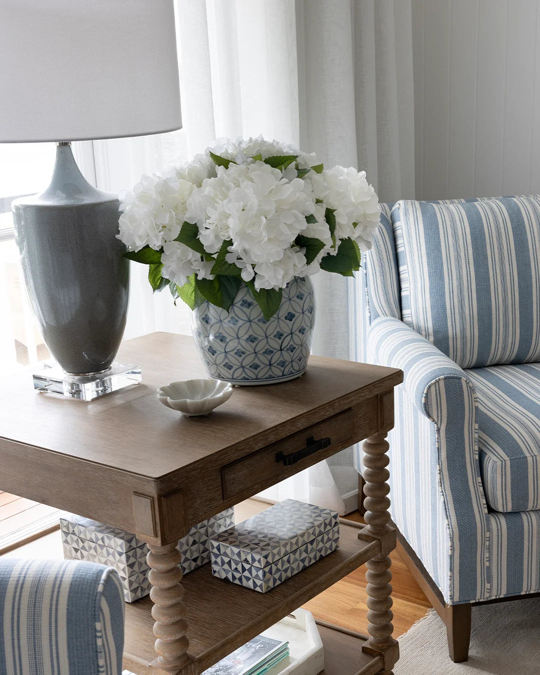 Side table styled with lage soft blue grey lamp, ginger jar filled with white faux hydrangea flowers, two blue triangle boxes on lower shelf. A striped armchair on either side of the side table.