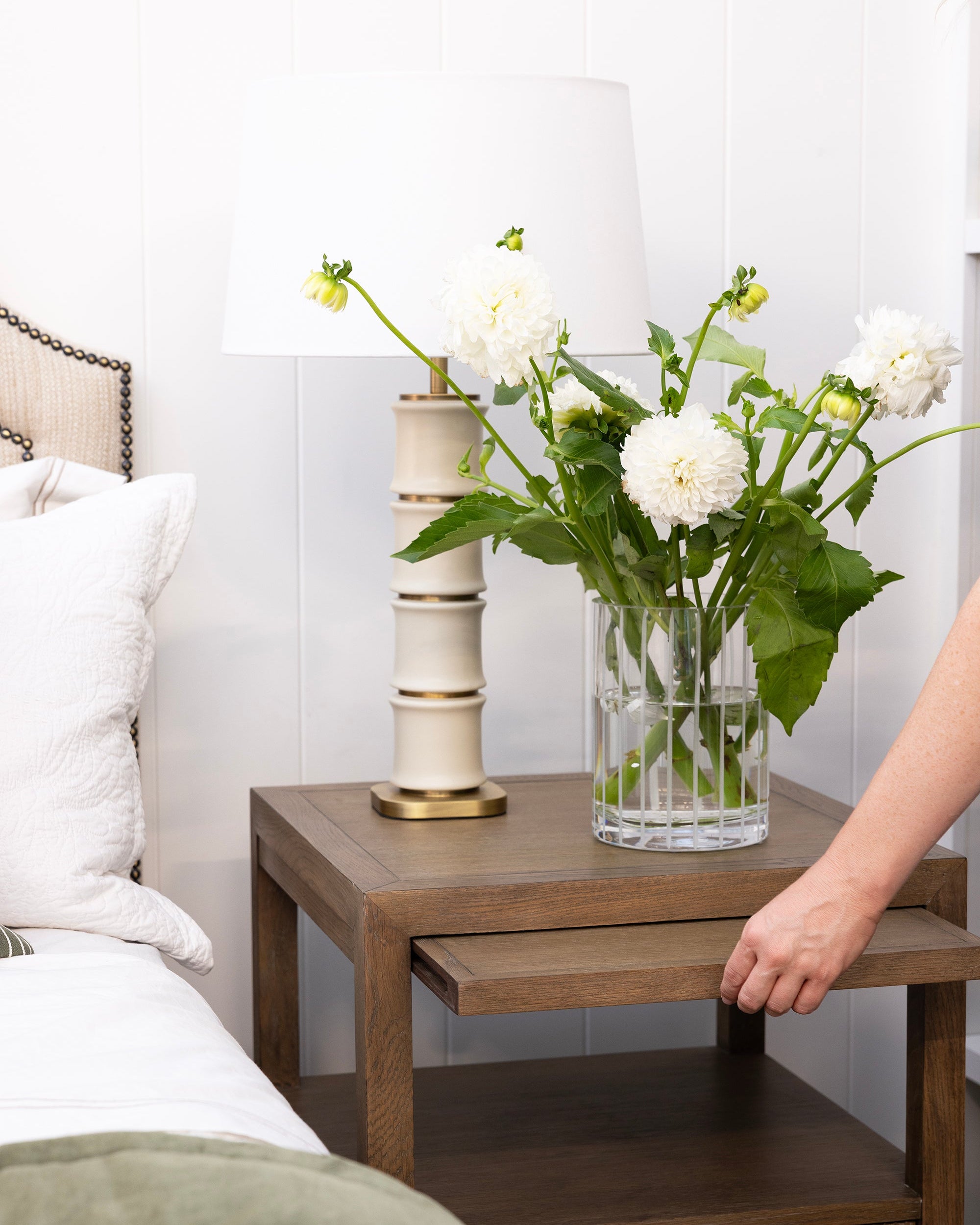 Wooden nightstand with a vase of flowers and a lamp next to a bed.