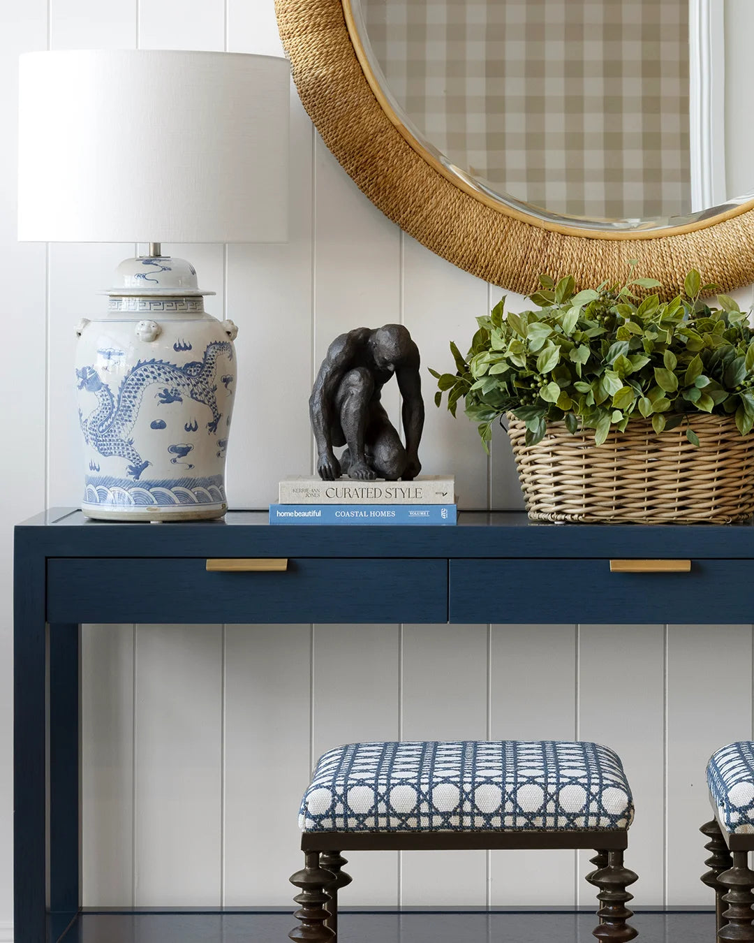 Navy console sitting in front of white wall. Woven round mirror above. Console is styled with blue and white ceramic lamp, crouching man on a stack of books and basket of faux greenery. A blue and white patterned ottoman sits in front.