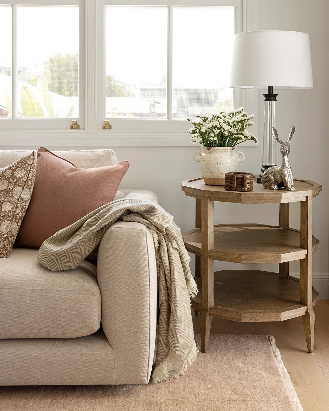 Geometric side table in oak sitting next to neutral sofa with beige throw and pink cushion. Side table is styled with a rustic vase of flowers, a set of dark rattan coasters, a silver bunny decorative object and a lamp.