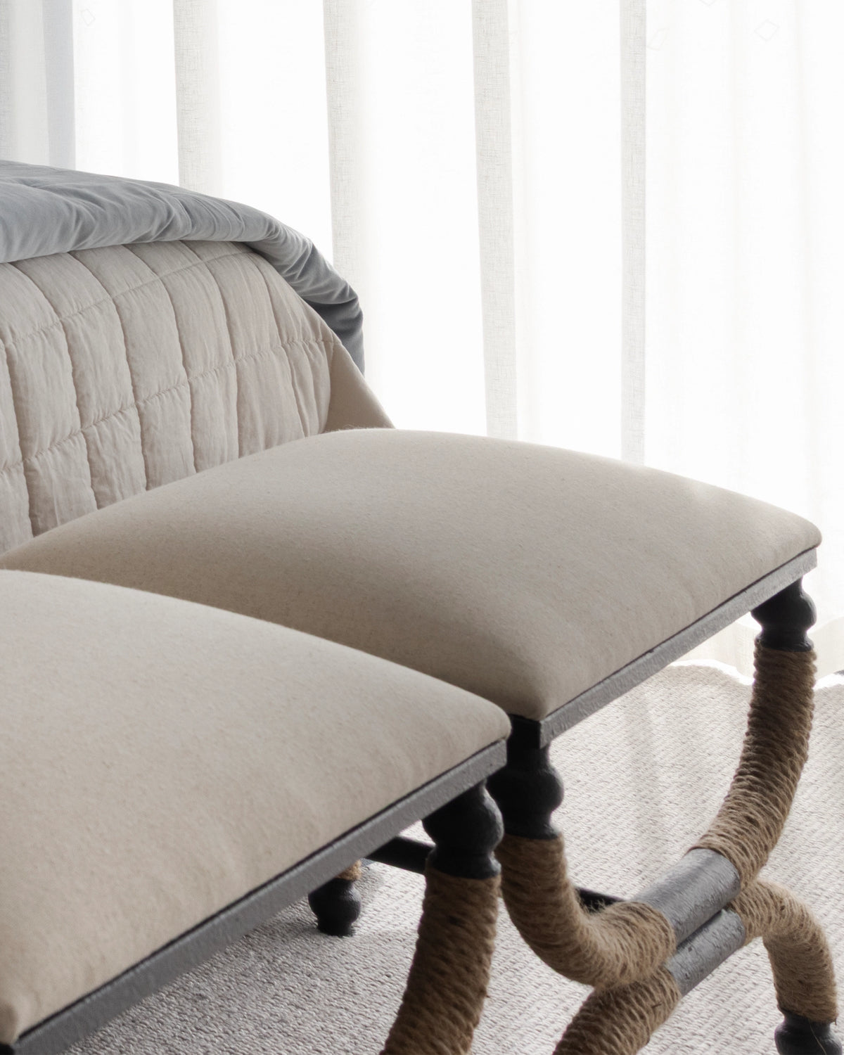 A close-up of upholstered bench and ottoman in warm beige fabric, with dark wooden legs wrapped in rope, set in a softly lit room with sheer curtains.