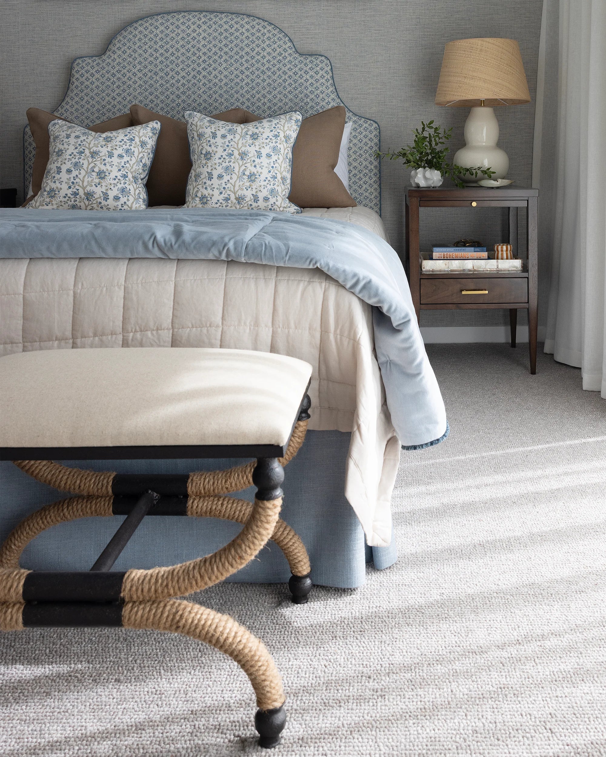 A styled bedroom scene featuring a bed with a light blue patterned upholstered headboard, beige and blue bedding, matching pillows, a rope-wrapped bench in the foreground, and a wooden nightstand with a lamp.