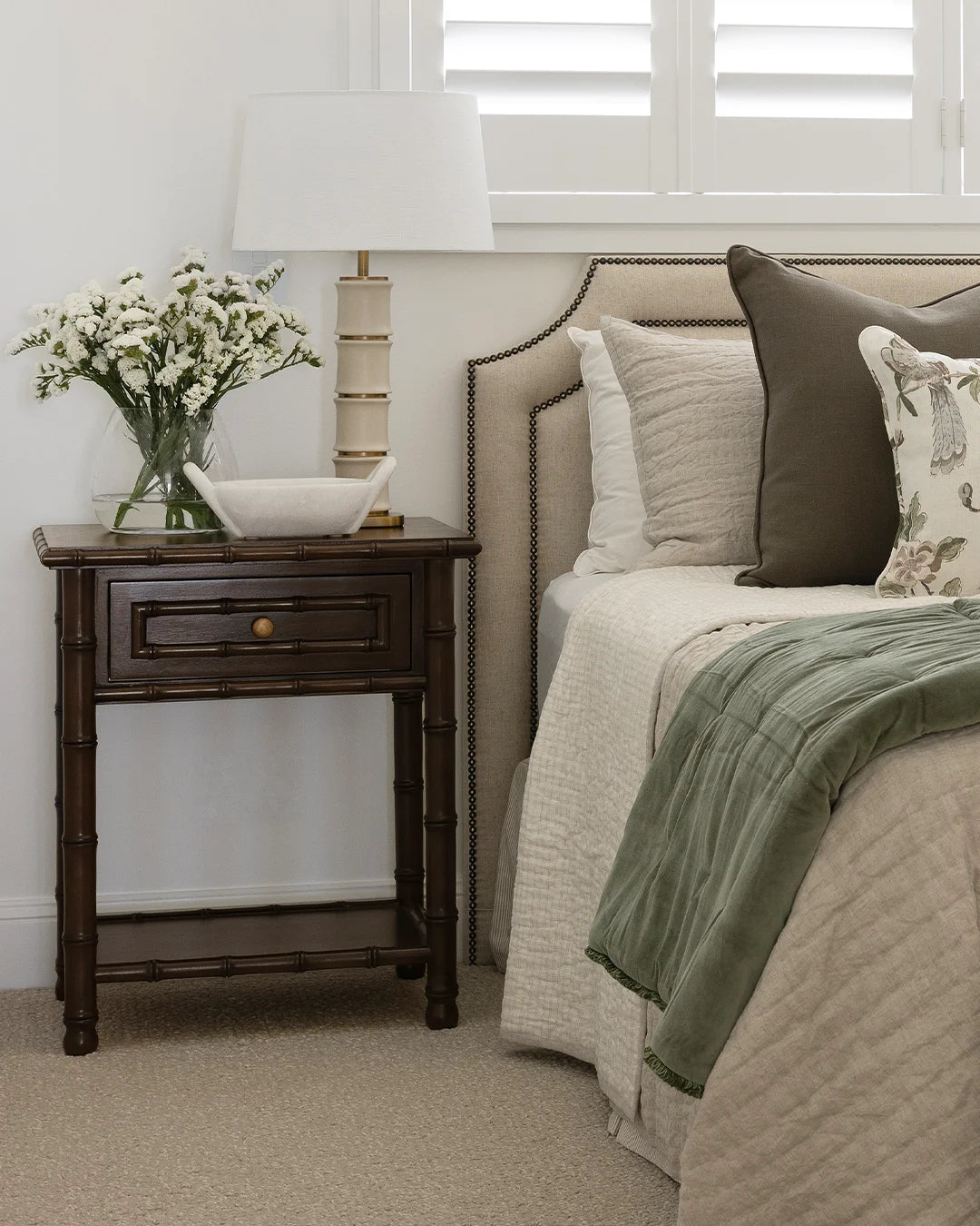 Faux bamboo timber bedside table styled with a vase of white flowers, carved marble bowl and a ceramic bamboo shaped lamp. Sitting next to a neutral made bed.