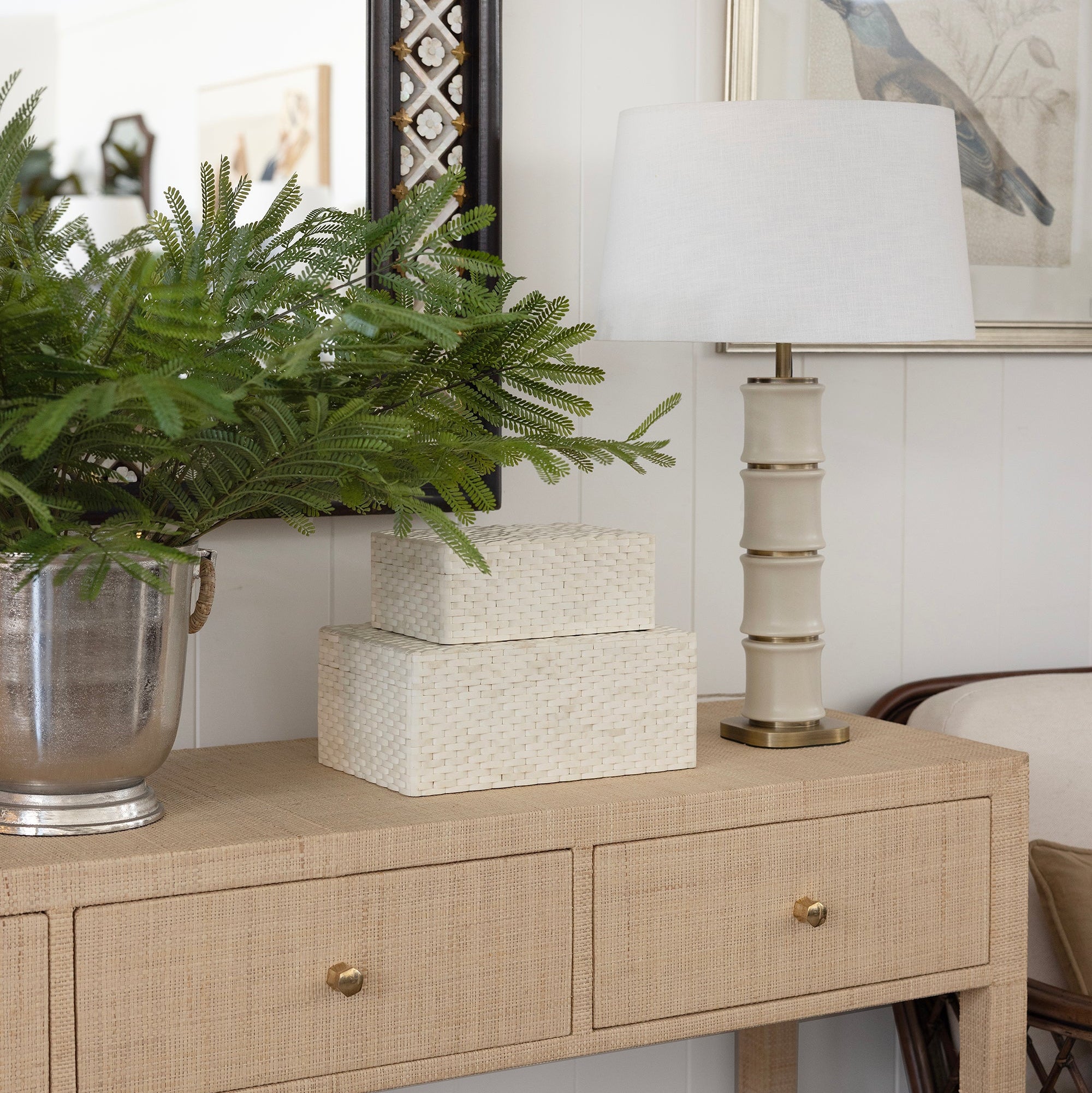 A raffia woven console table in a styled room setting, decorated with a green fern in a silver pot, two stacked white textured boxes, and a cream-colored table lamp with a white shade.