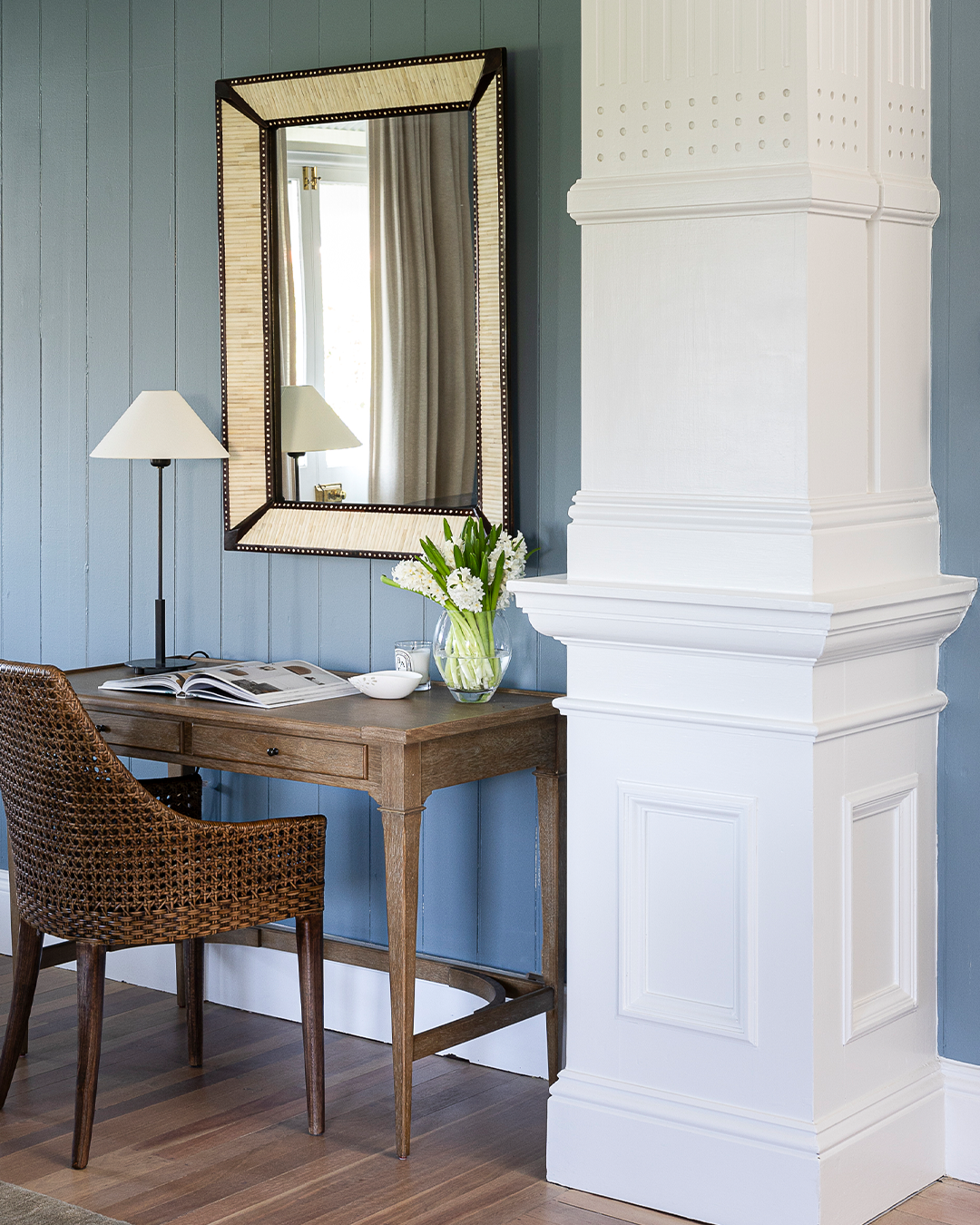 A styled interior corner featuring a dark wood writing desk with a woven wicker chair, a tall rectangular mirror with a black and gold frame, a floor lamp, and a vase of flowers against a blue paneled wall next to a white architectural column.
